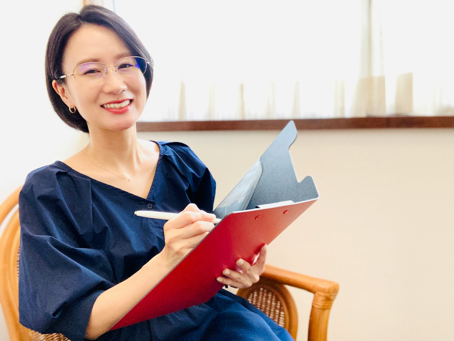 Seorang wanita tersenyum mengenakan gaun biru memegang clipboard merah di ruangan terang