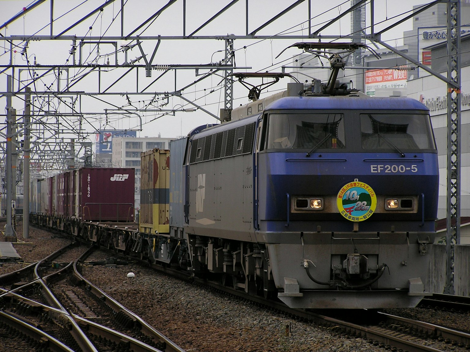 Un tren de carga azul moviéndose a lo largo de las vías con un paisaje urbano al fondo