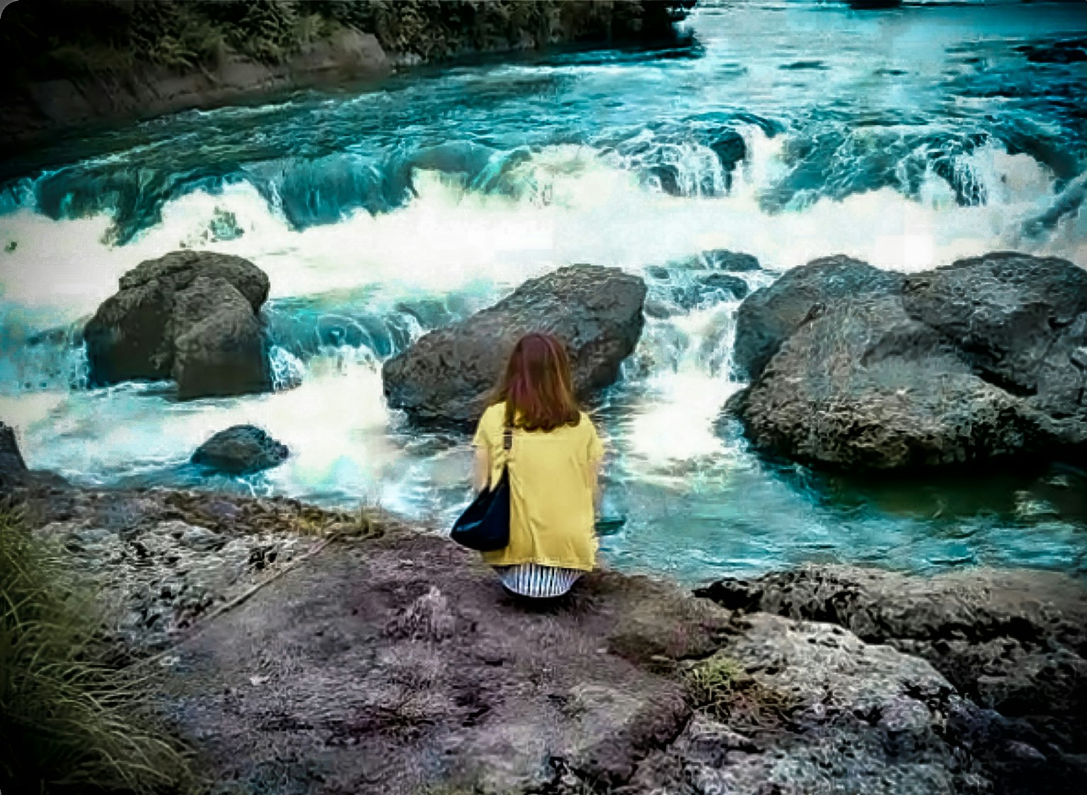 Una donna in cardigan giallo è seduta su rocce vicino a un fiume in piena