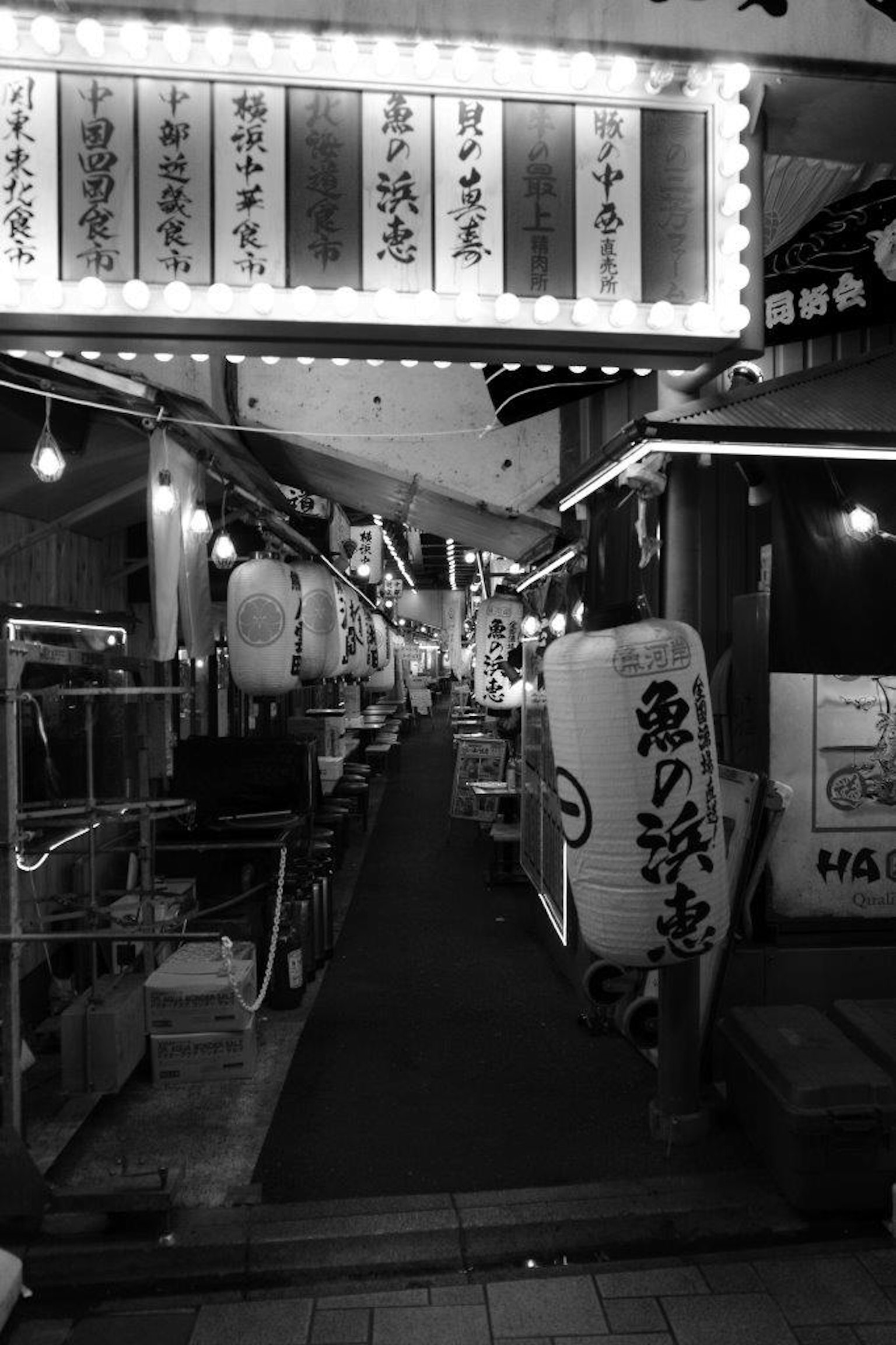 Black and white image of a narrow street lined with lanterns and restaurant signs
