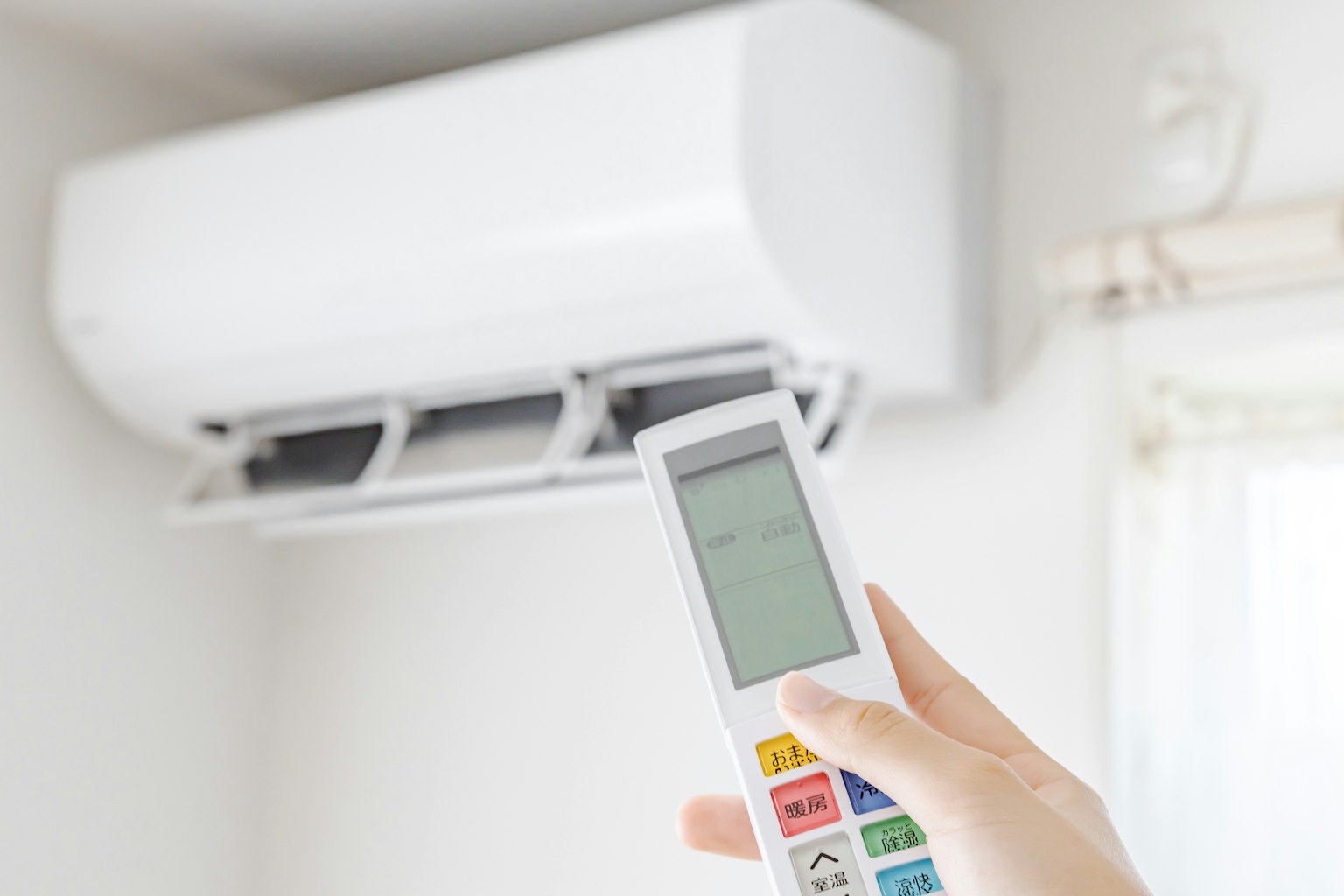 Tangan memegang remote AC di depan unit AC putih