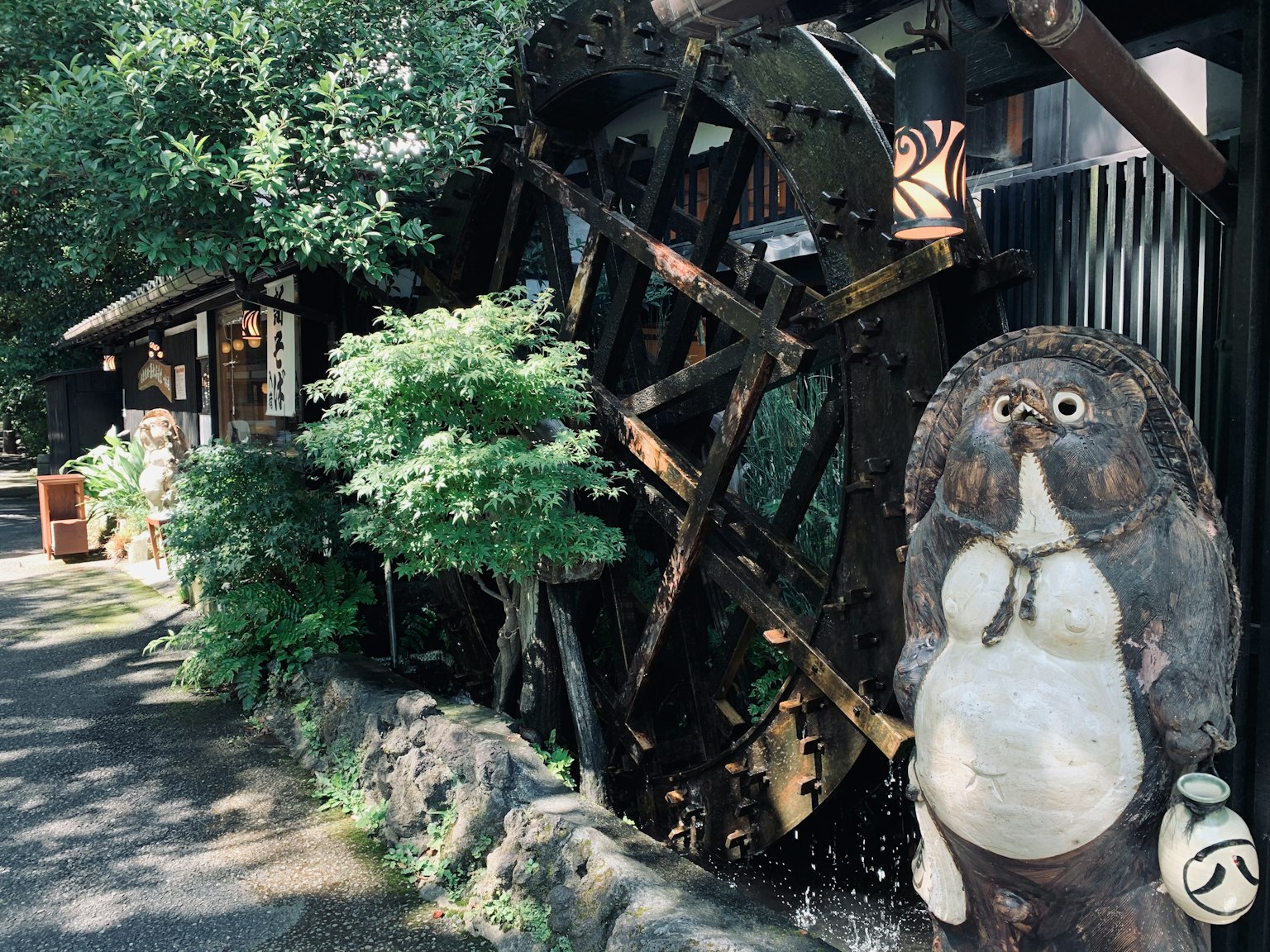A tanuki statue near a water wheel with lush greenery