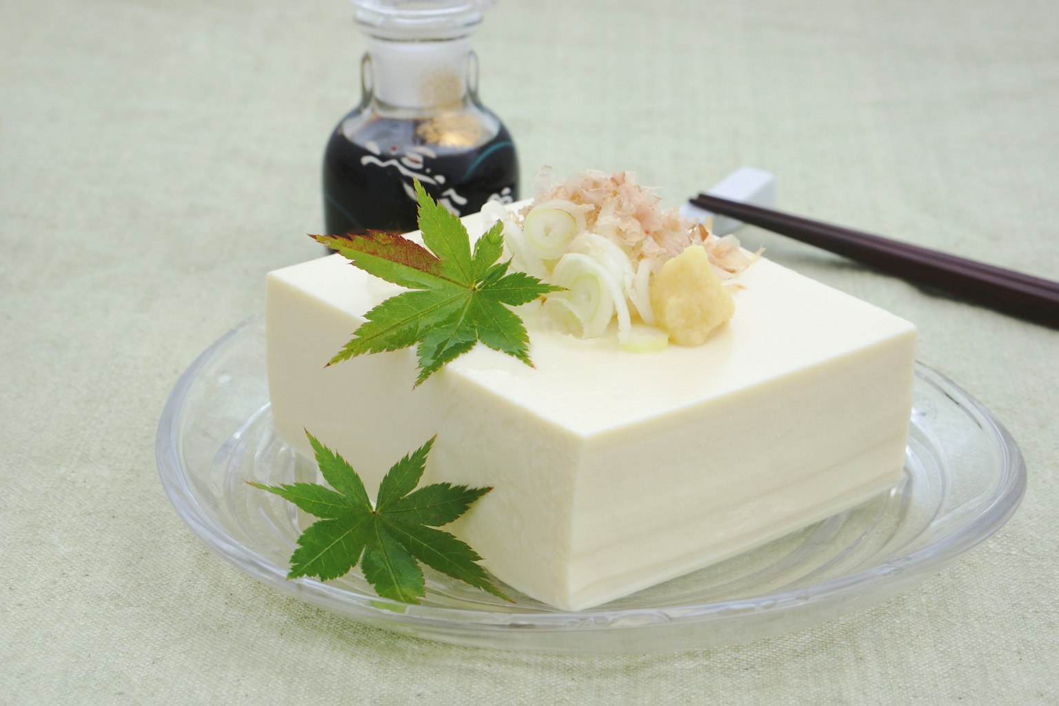 Un bloc di tofu guarnito con zenzero e scaglie di tonnetto accanto a una bottiglia di salsa di soia e bacchette