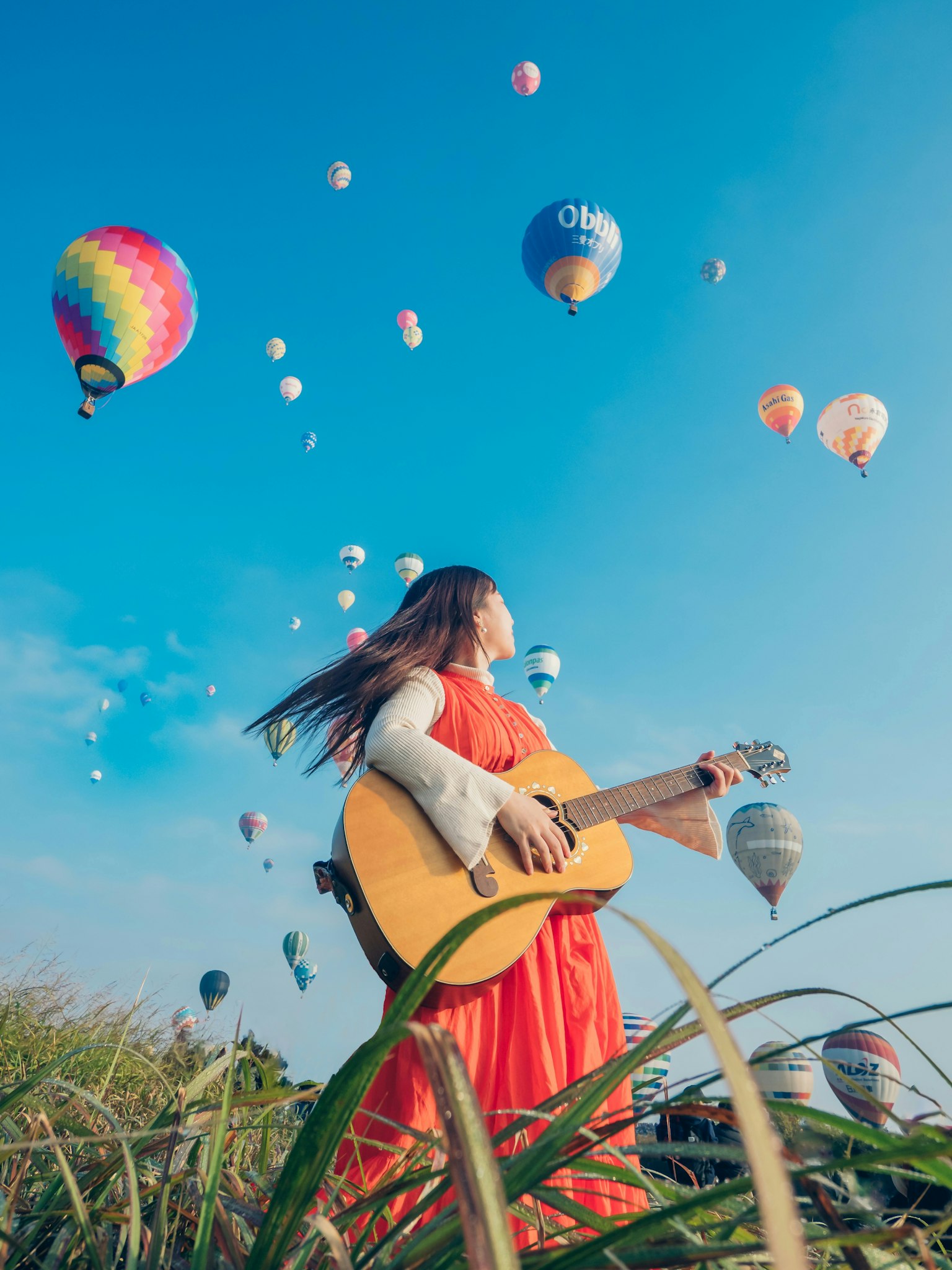 Una mujer sosteniendo una guitarra bajo un cielo azul con globos aerostáticos coloridos