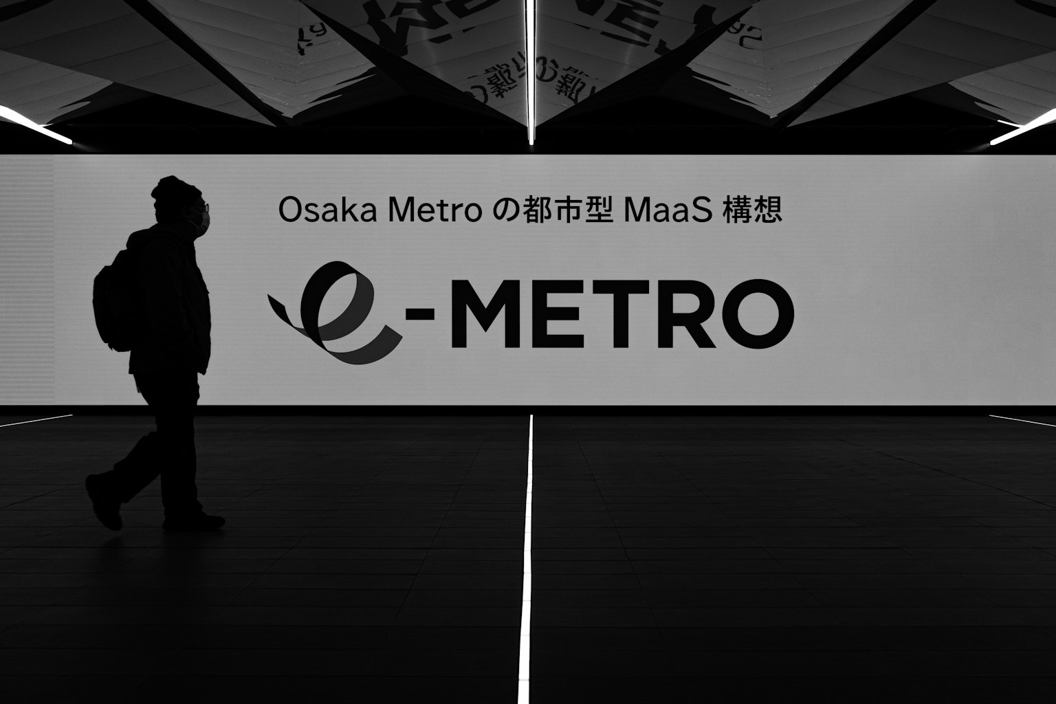 เงาของคนที่เดินผ่านโลโก้ e-METRO ของโอซาก้าเมโทร