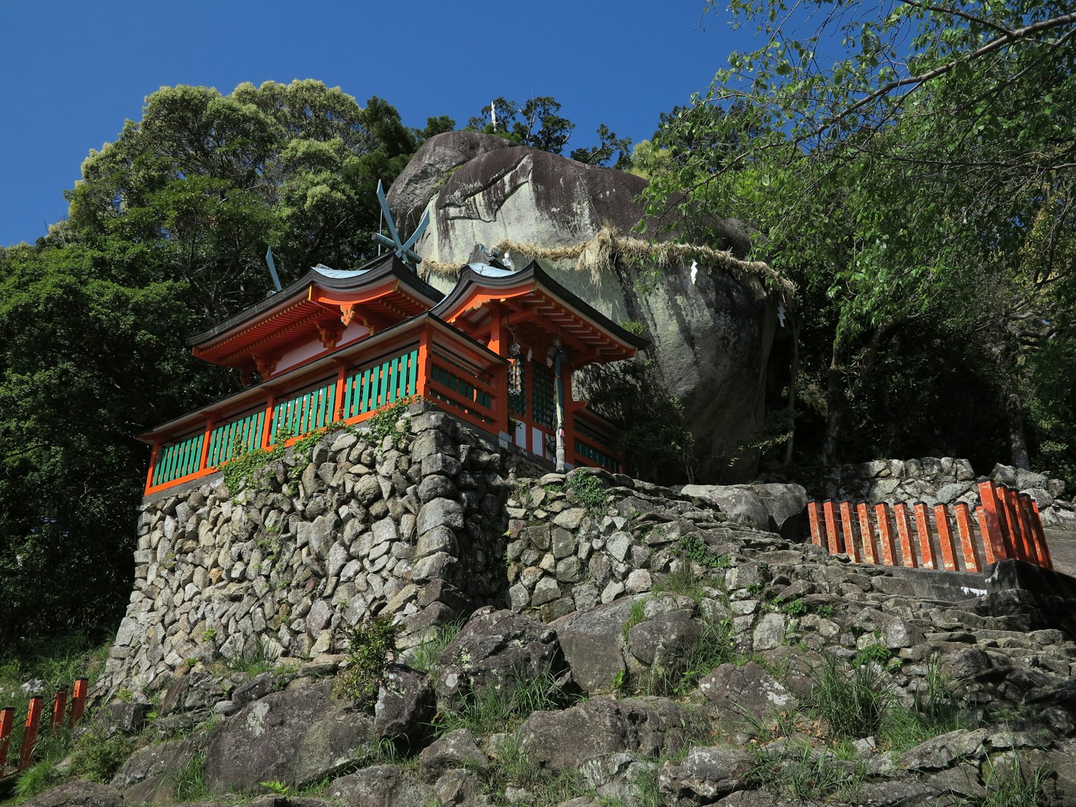 一座橙色和绿色的神社建筑坐落在被绿色植物环绕的岩石山坡上