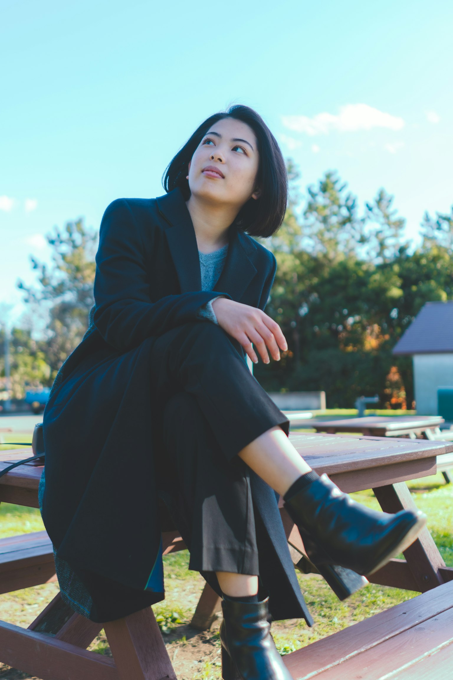 一名女性坐在公园长椅上，穿着黑色外套和靴子，背景是蓝天