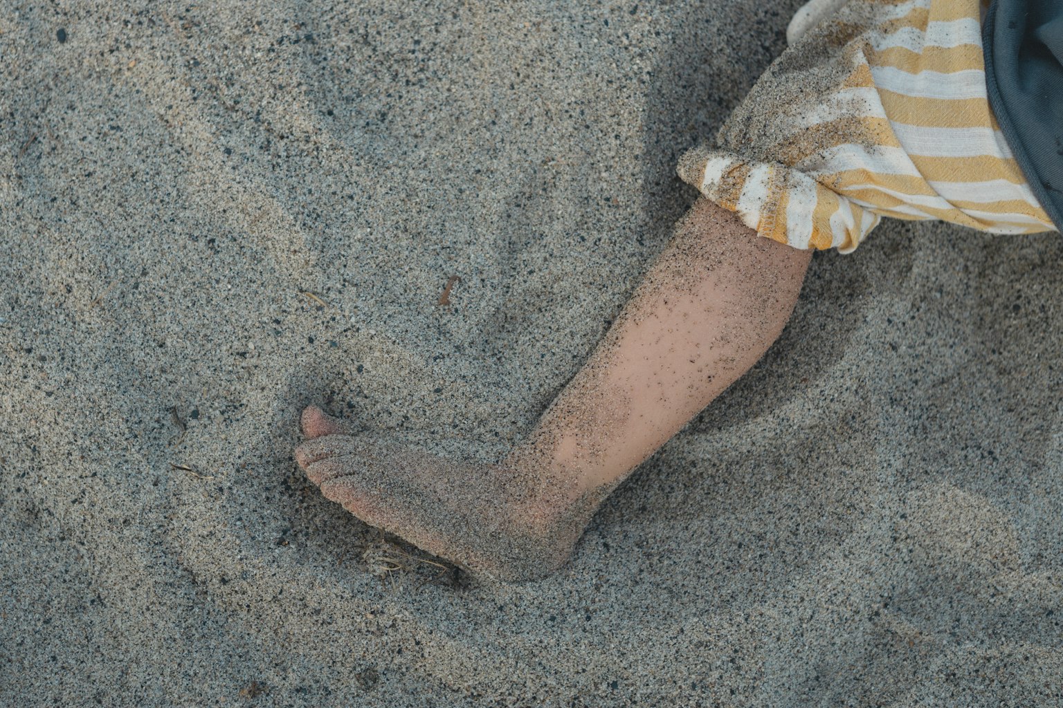Kaki yang sebagian tertimbun pasir dengan anak yang mengenakan celana bergaris