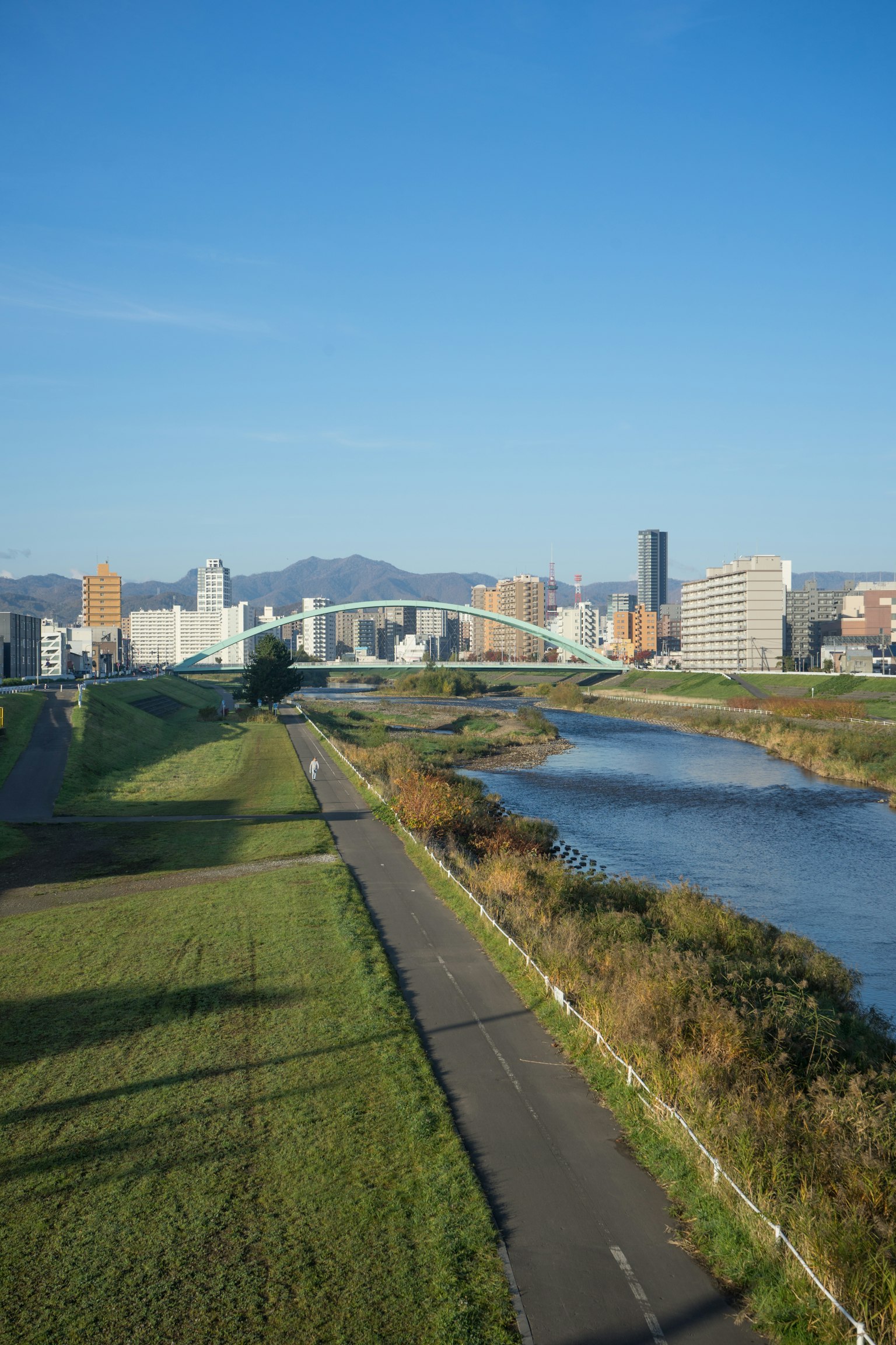 Cityscape with a river under a blue sky featuring a bike path and a bridge