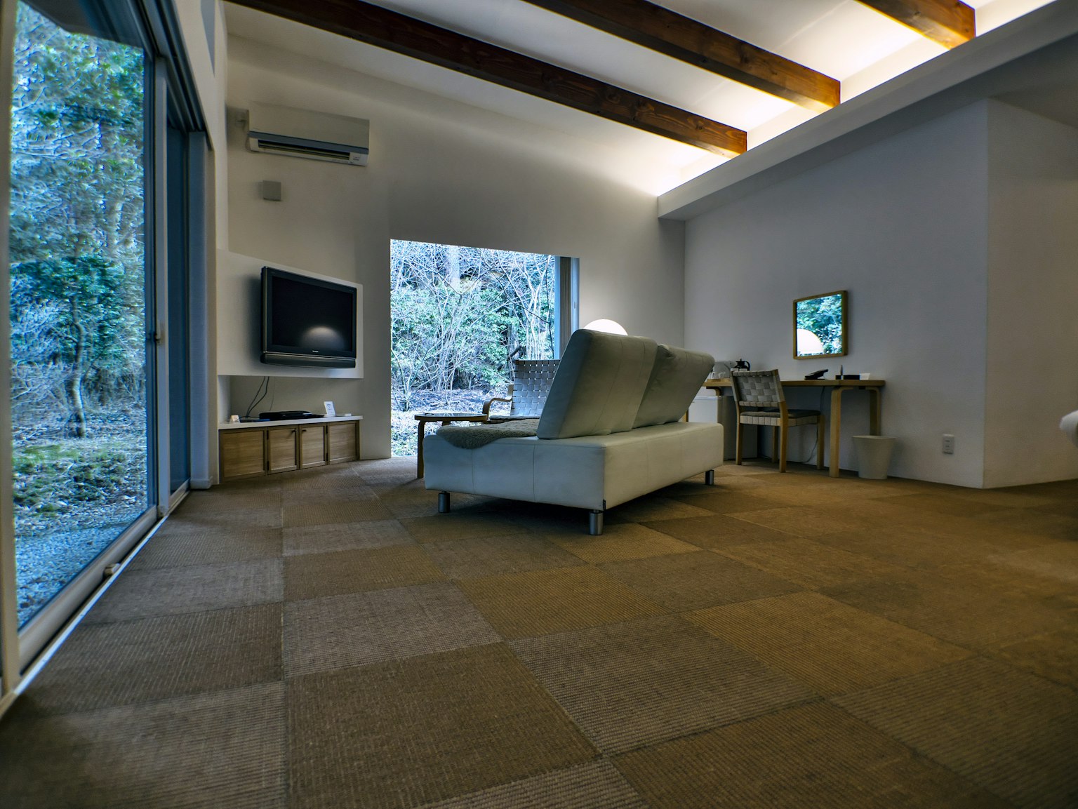 Interior de sala moderna con grandes ventanas que dejan entrar luz natural vigas de madera y disposición simple de sofá y escritorio