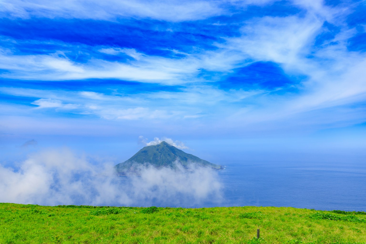 A small island surrounded by blue sky and clouds above a green meadow
