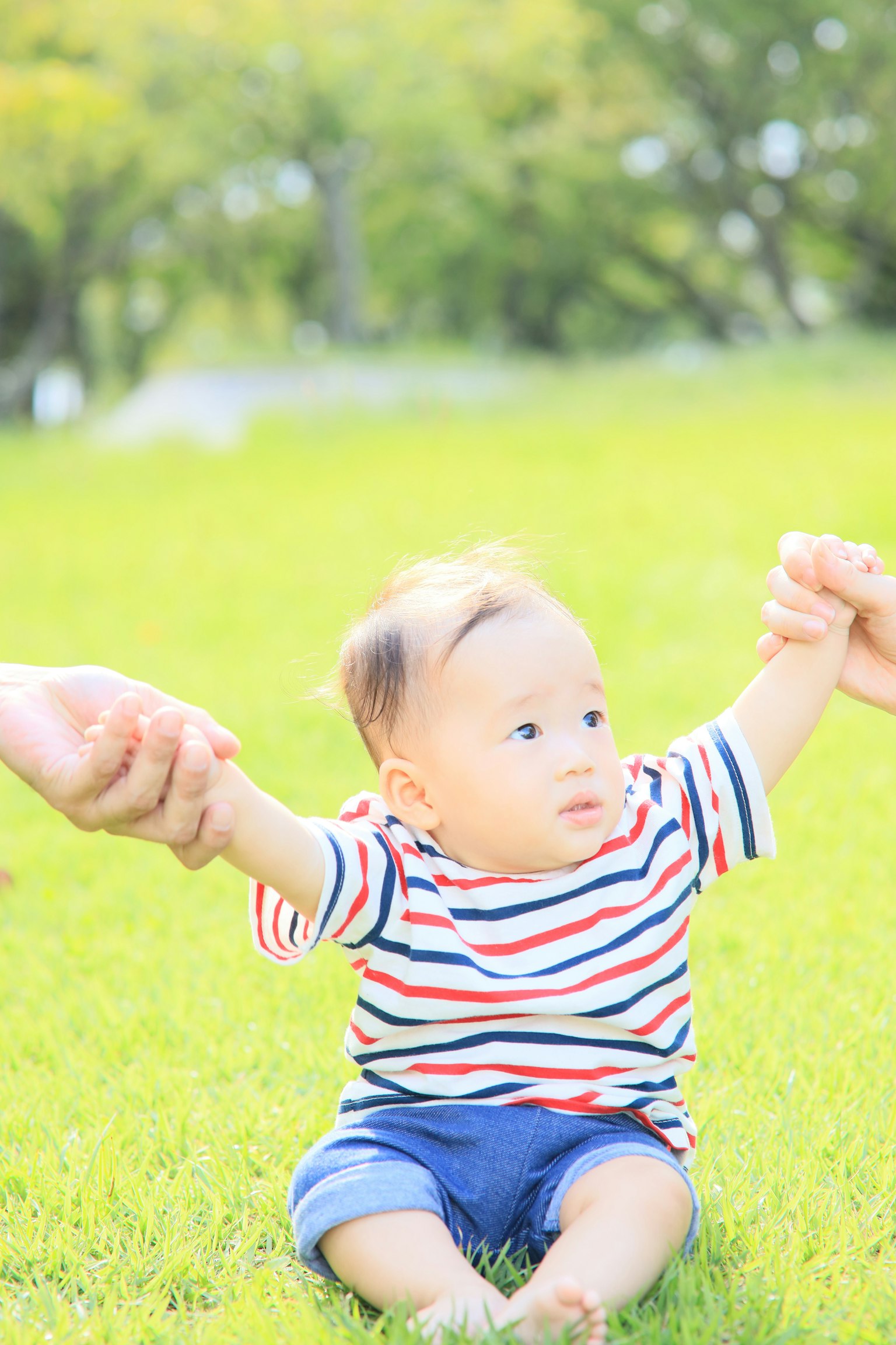 Seorang bayi duduk di rumput memegang tangan orang dewasa