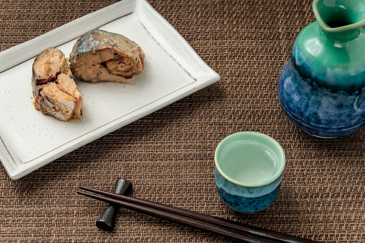Grilled fish on a white plate with a blue sake bottle and small cup on a textured table