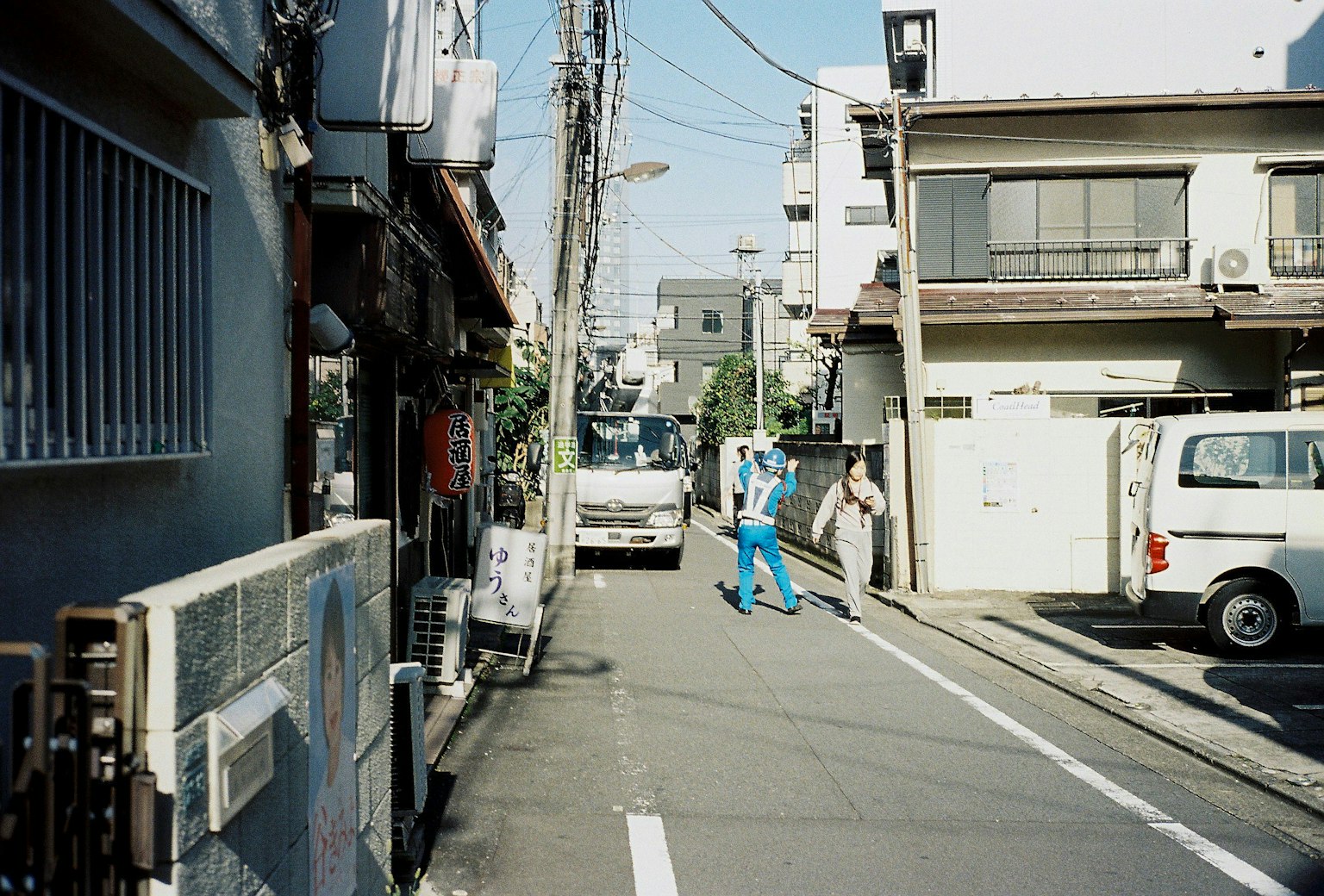 Narrow street scene with residential buildings and shops featuring a person in blue work clothes walking