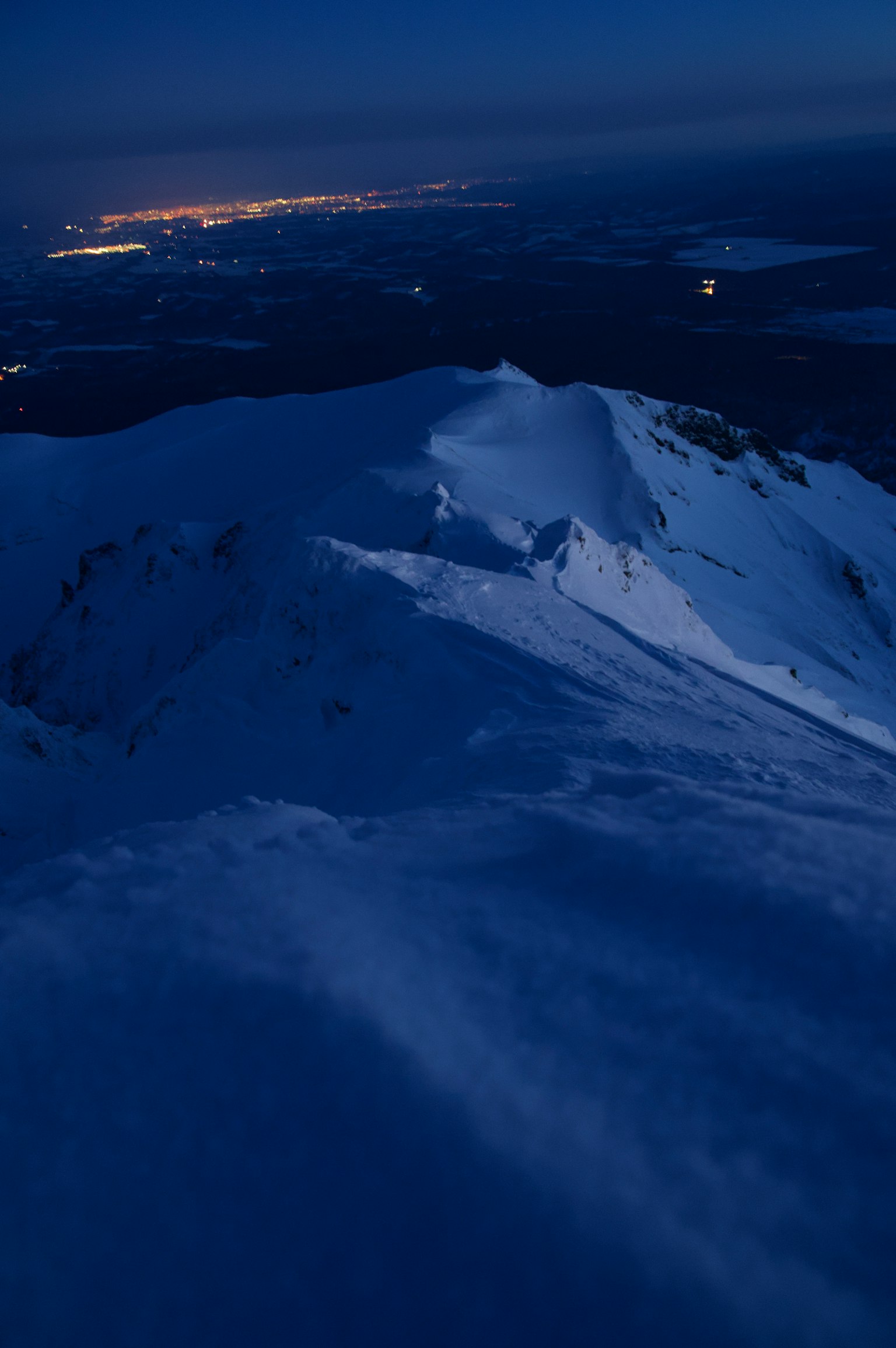 Snow-covered mountain peak at night with distant city lights