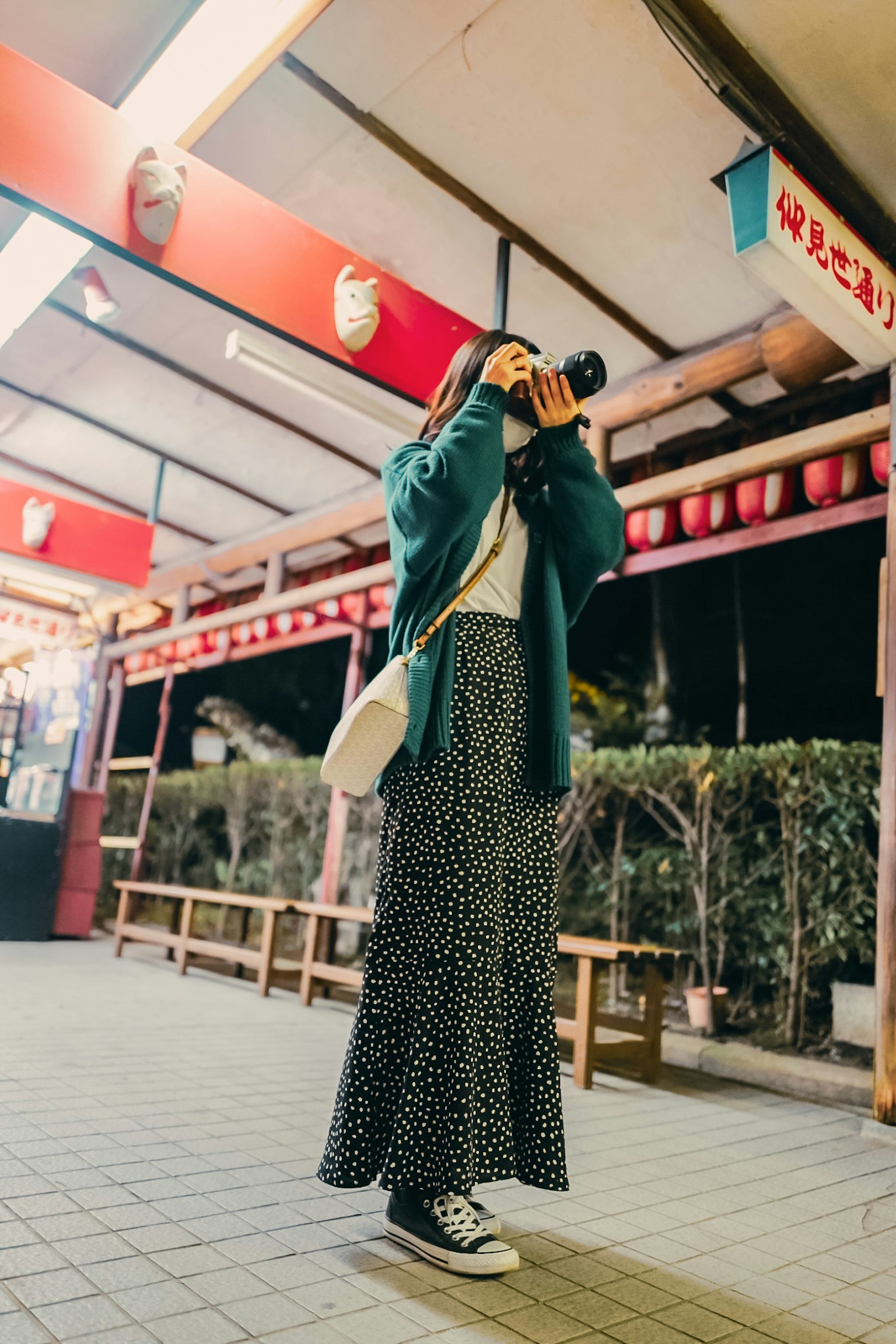 Femme tenant un appareil photo dans la rue la nuit portant une veste verte et une jupe à motifs entourée de lanternes rouges