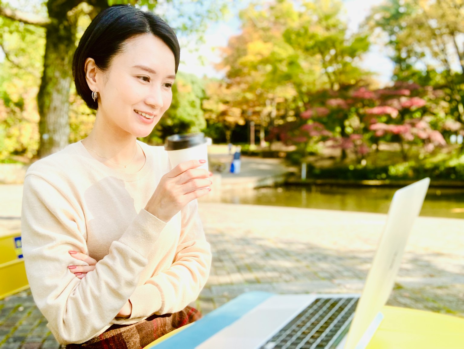 Frau, die lächelt, während sie einen Kaffee hält und in einem Park einen Laptop benutzt