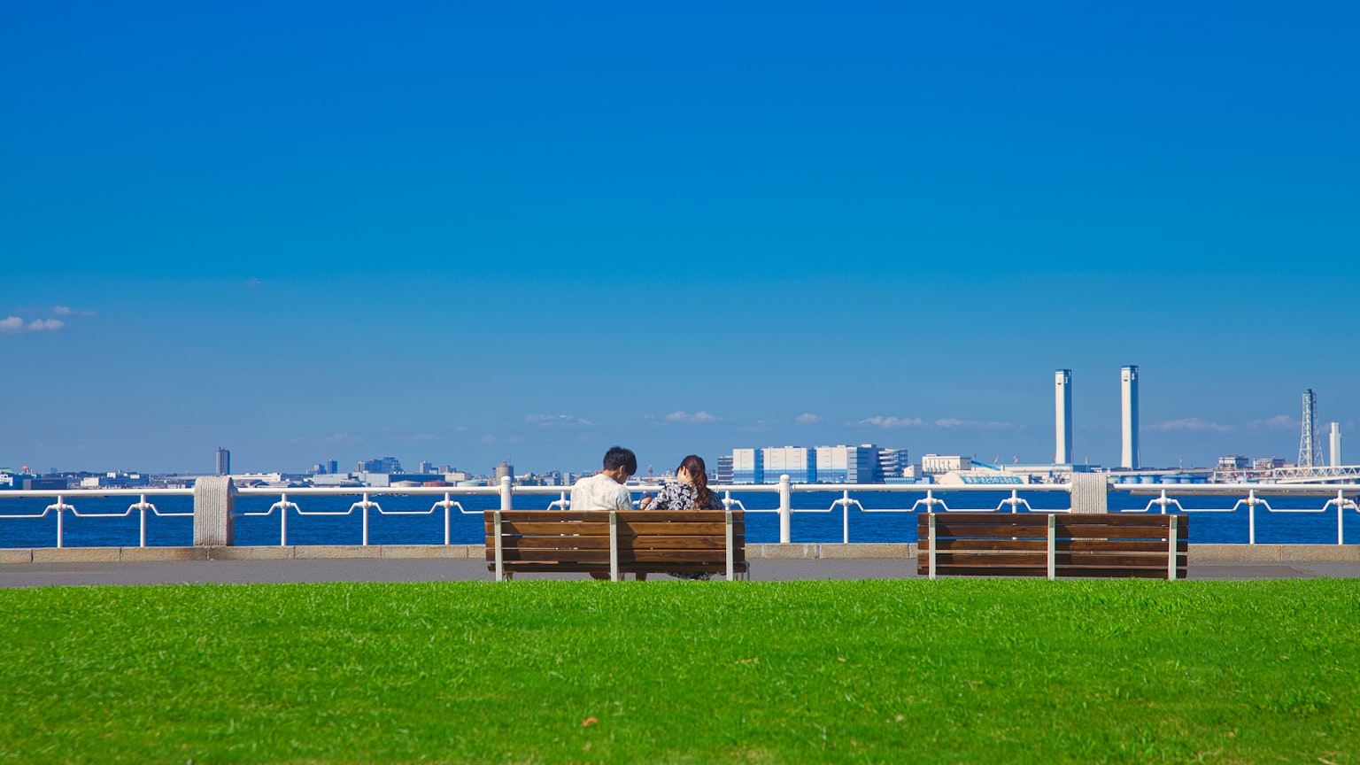 Dos personas sentadas en bancos bajo un cielo azul con vista al agua