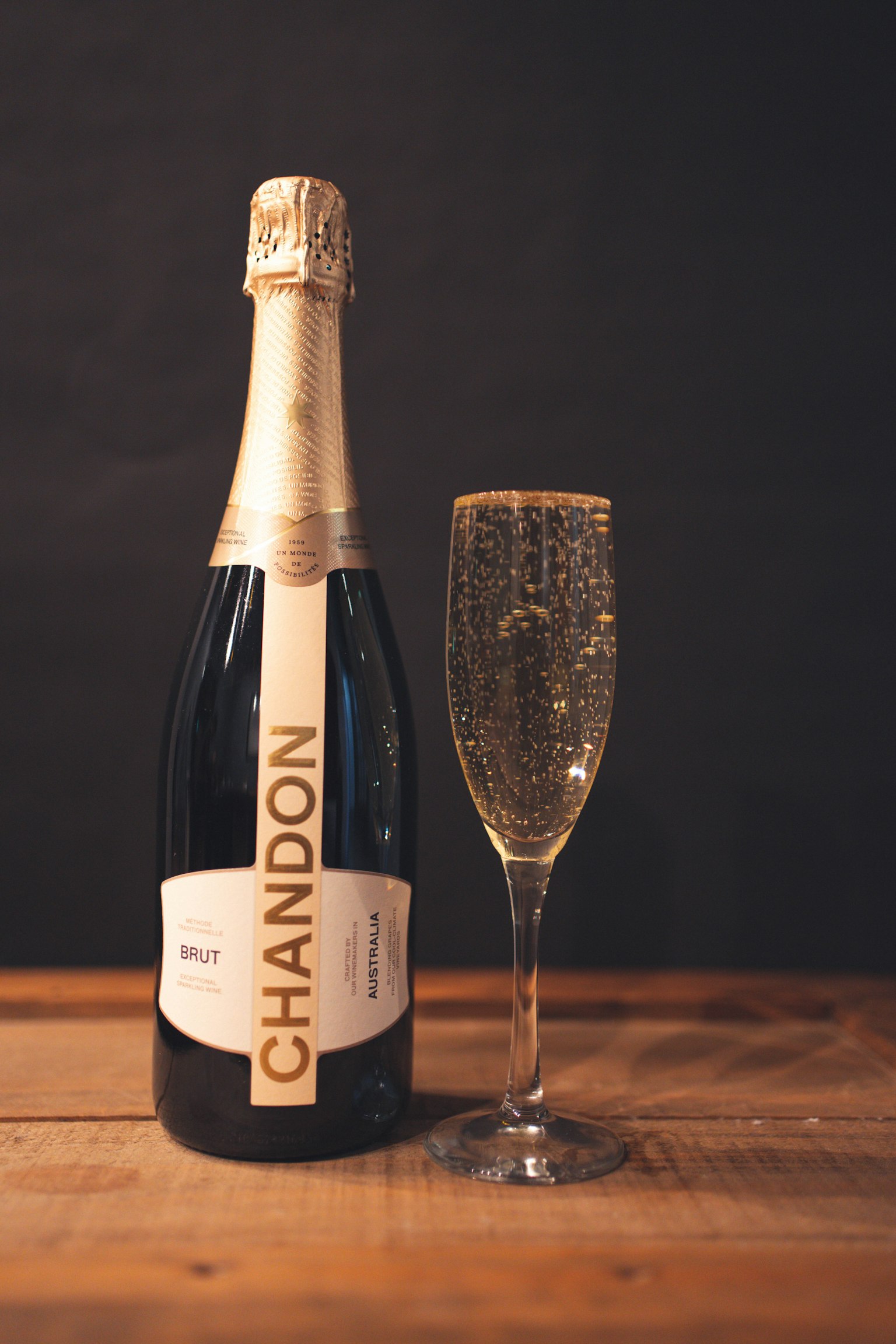 Chai rượu sâm panh Chandon bên cạnh ly sâm panh đầy trên bàn gỗ