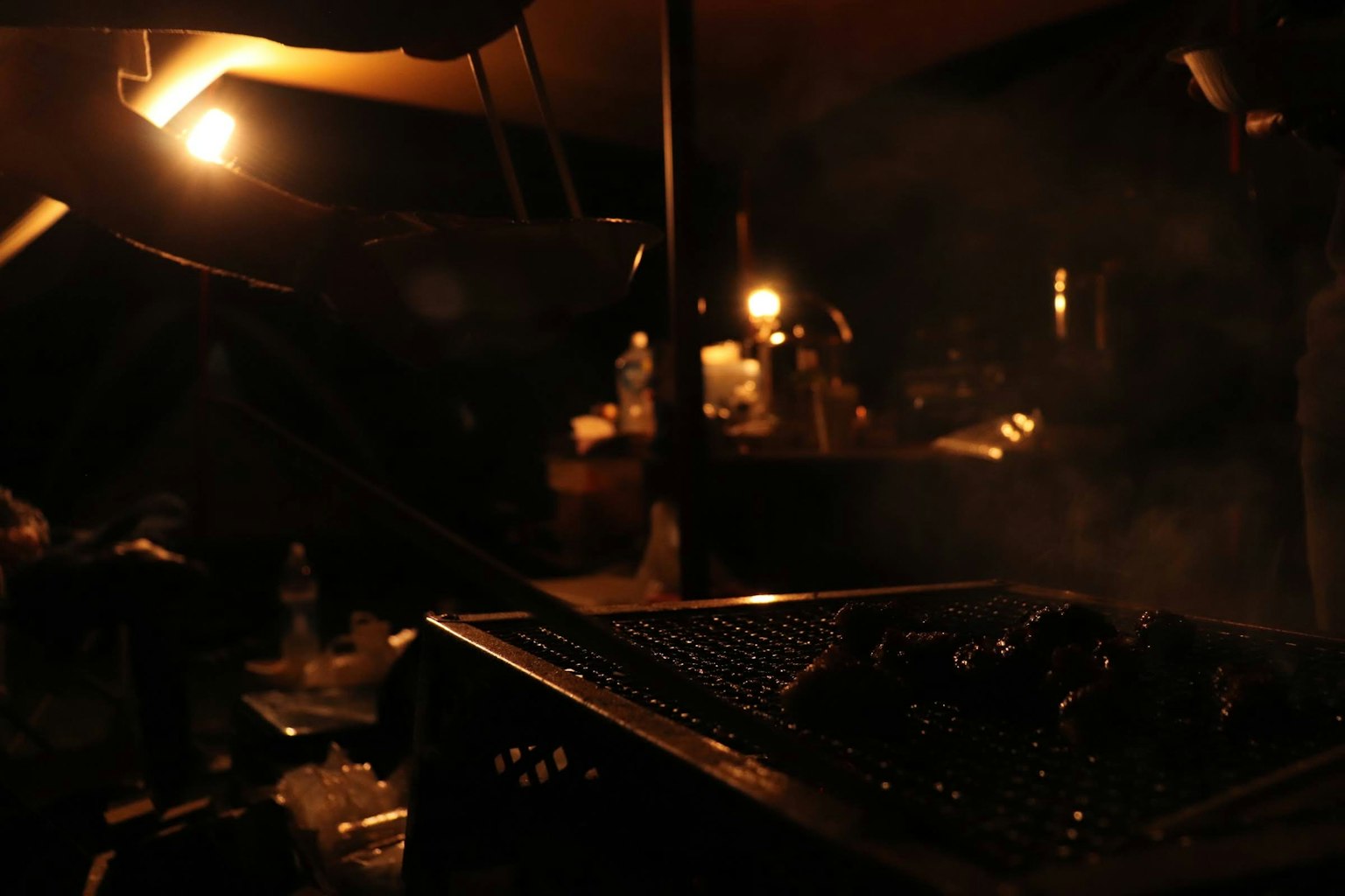 Grilled meat emitting smoke in a dark setting with dim surrounding lights