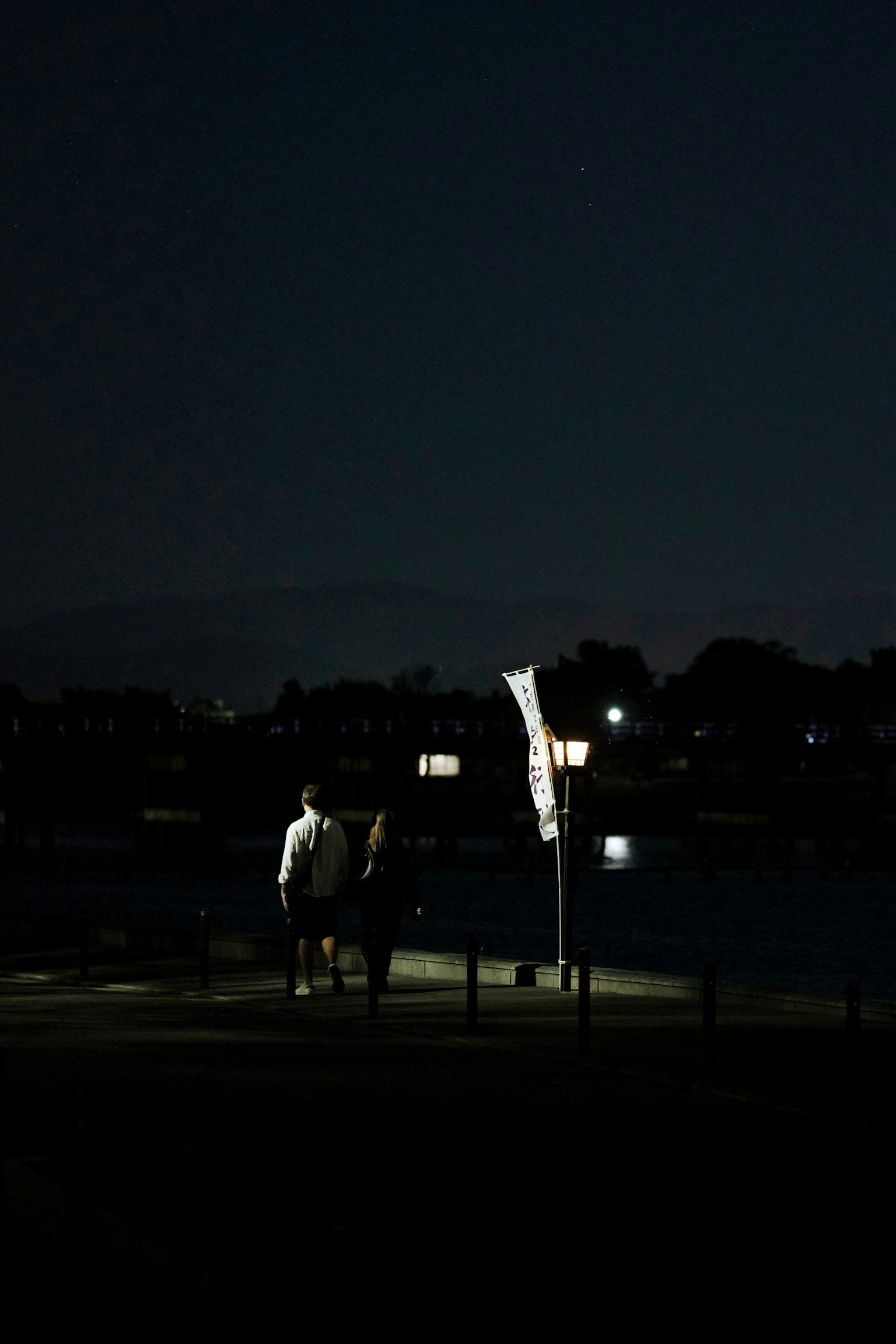 Dua sosok berdiri di tepi sungai di malam hari dengan pencahayaan lampu jalan