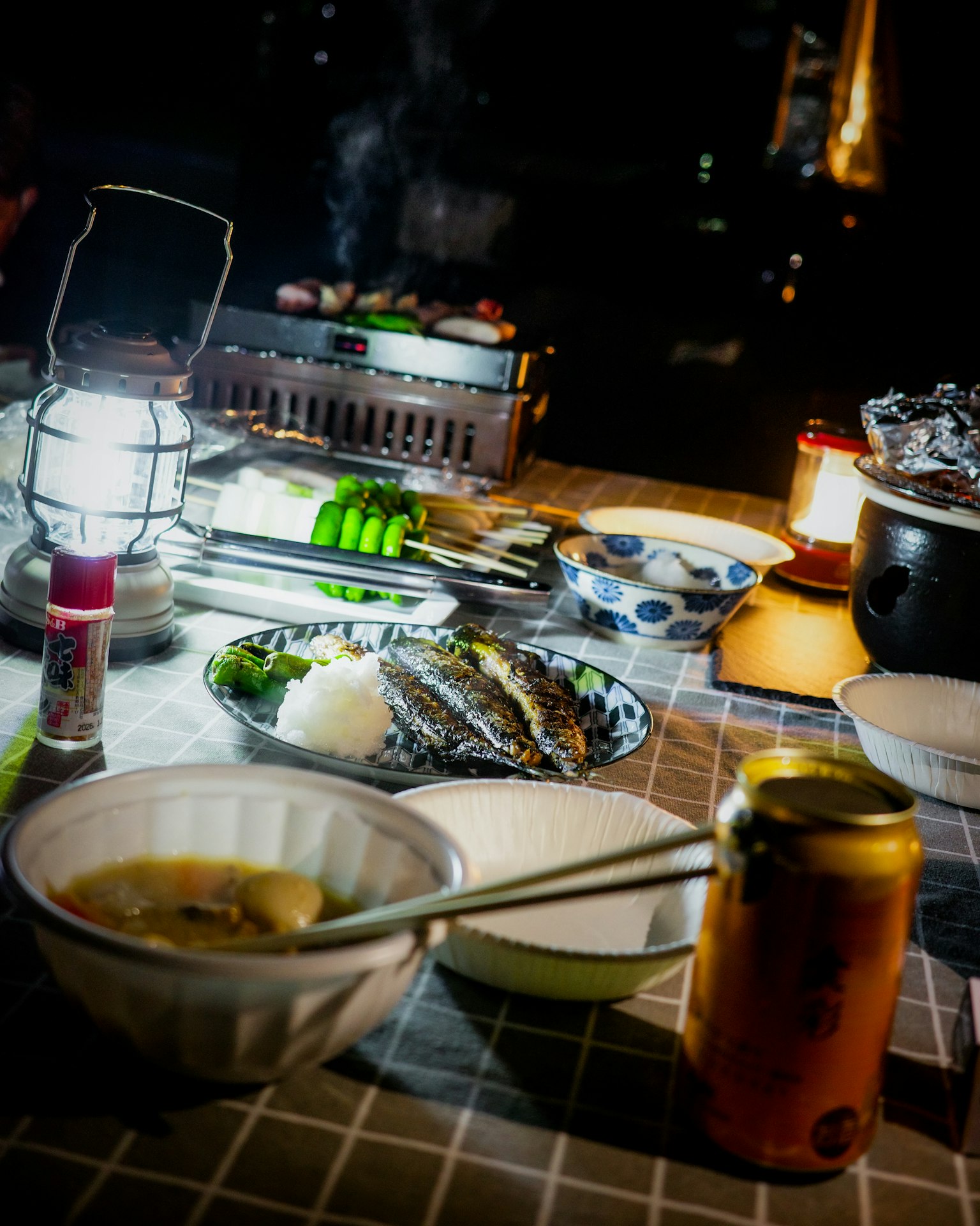 Outdoor barbecue scene at night featuring various dishes and drinks on a lit table