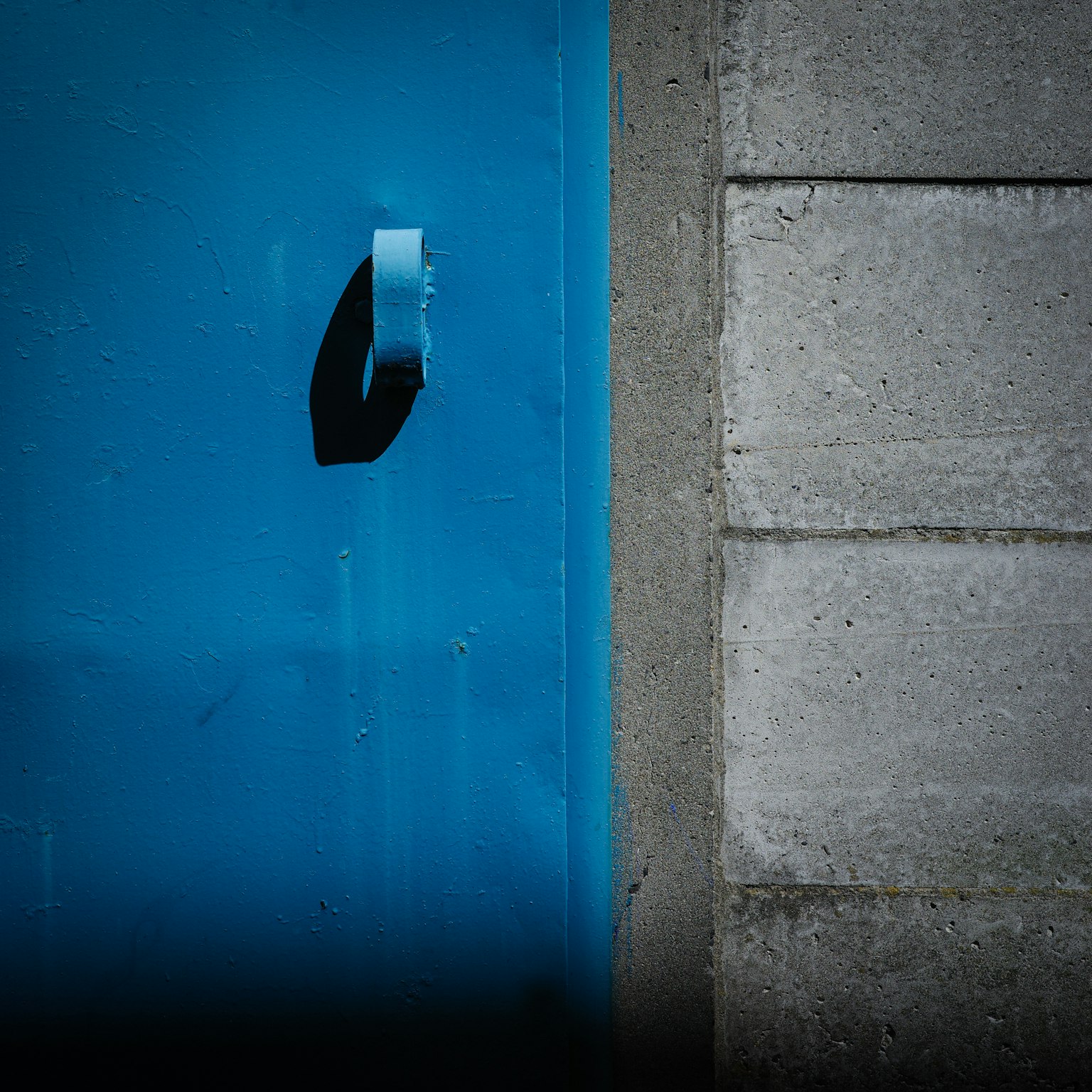 Vista parziale di una porta blu con una maniglia nera accanto a un muro di cemento