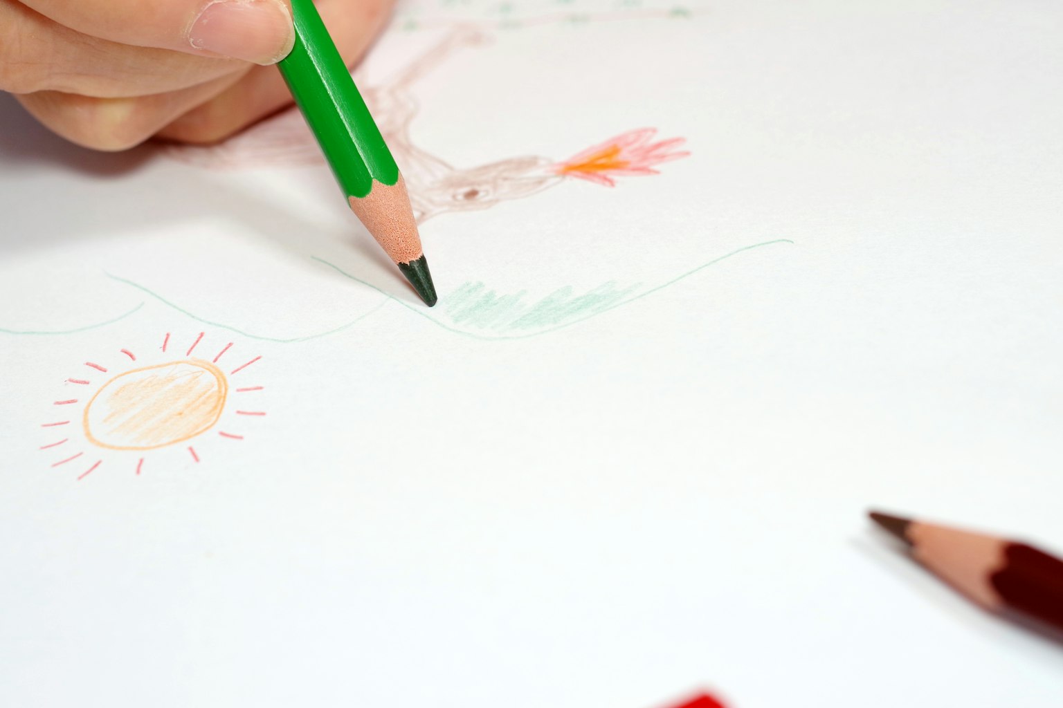 Mano de un niño sosteniendo un lápiz de color verde dibujando en papel con bocetos coloridos