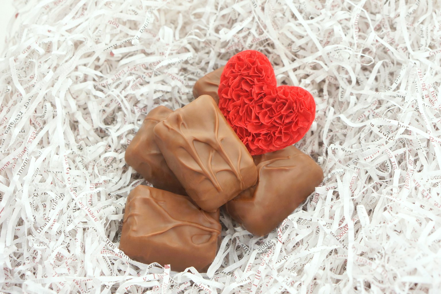 Un objet en forme de cœur rouge et des morceaux de chocolat placés sur du papier déchiqueté blanc