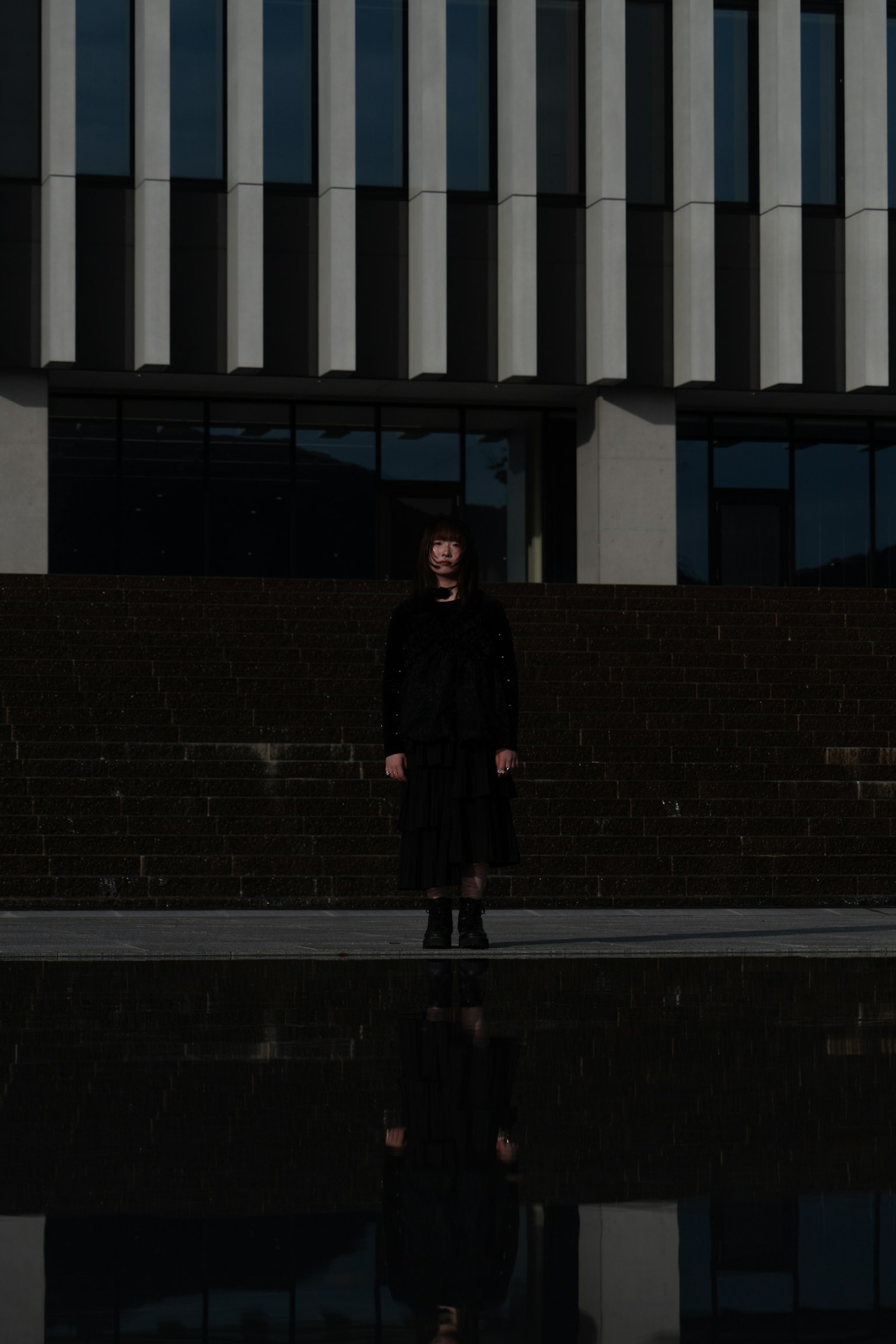 暗い服を着た人物が現代的な建物の前に立っている 反射が水面に映っている