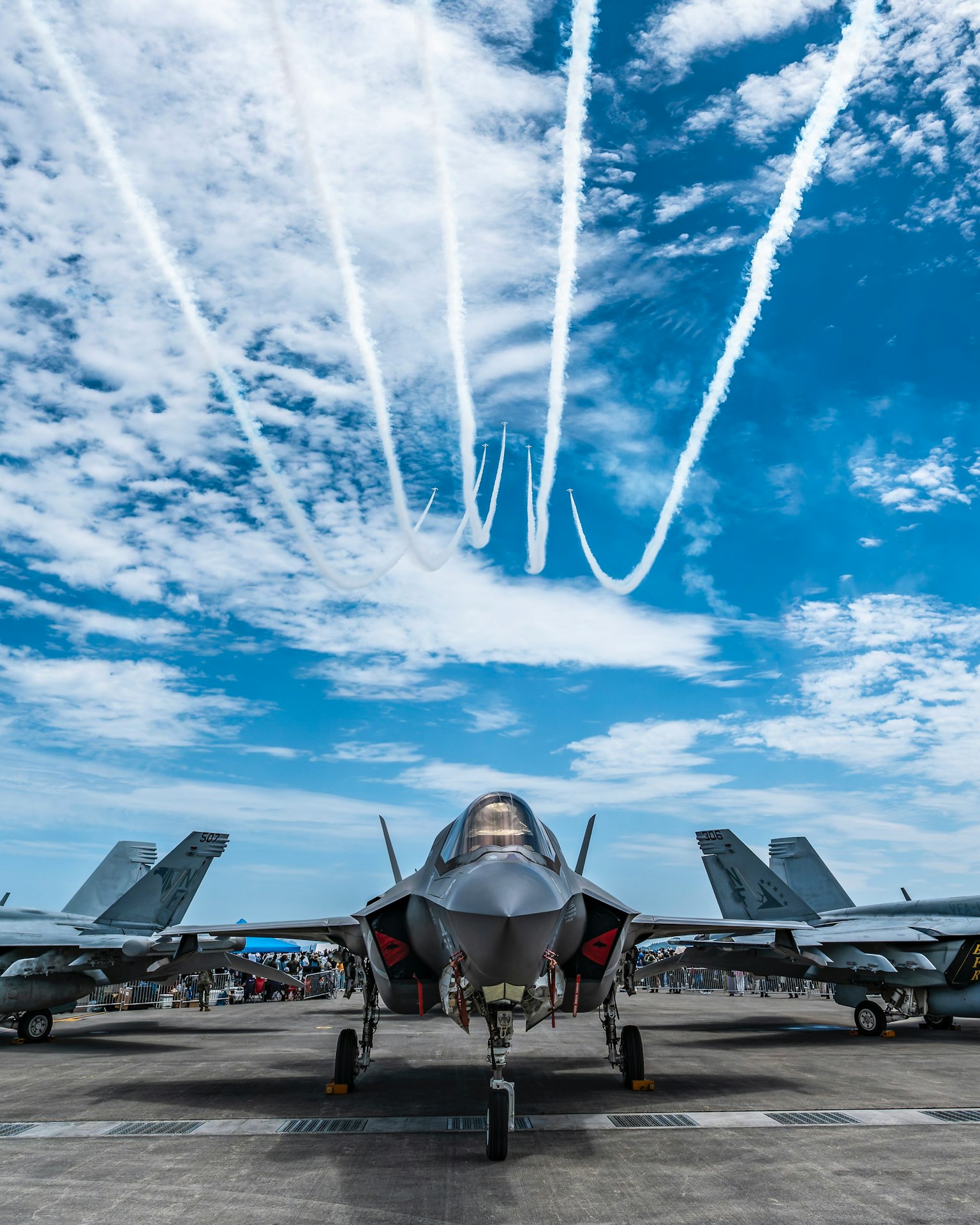 เครื่องบินขับไล่ F-35 ตั้งอยู่ด้านหน้าภายใต้ท้องฟ้าสีฟ้าพร้อมรอยทางอากาศในพื้นหลัง