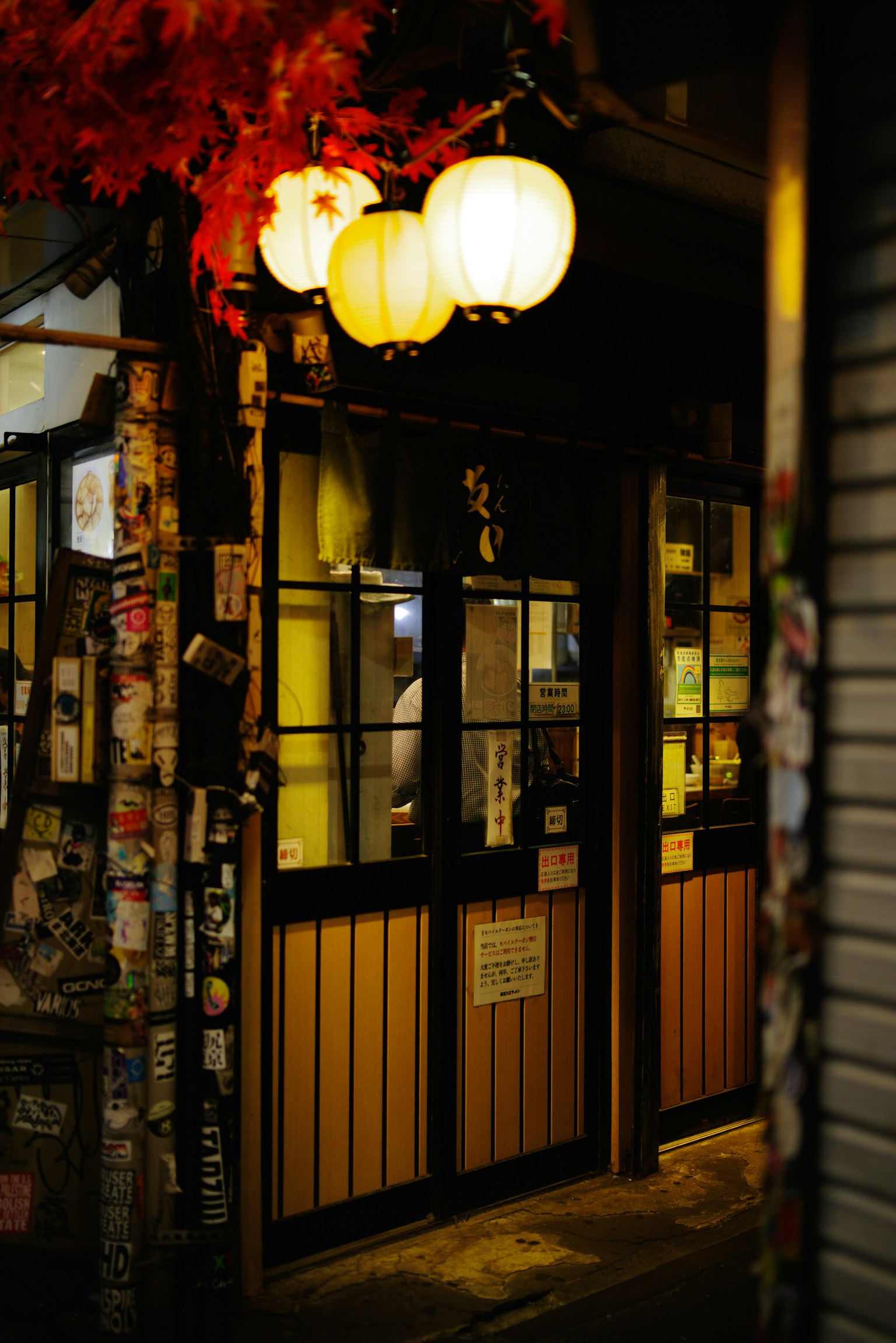 Entrance of a Japanese izakaya at night with hanging lanterns and colorful stickers