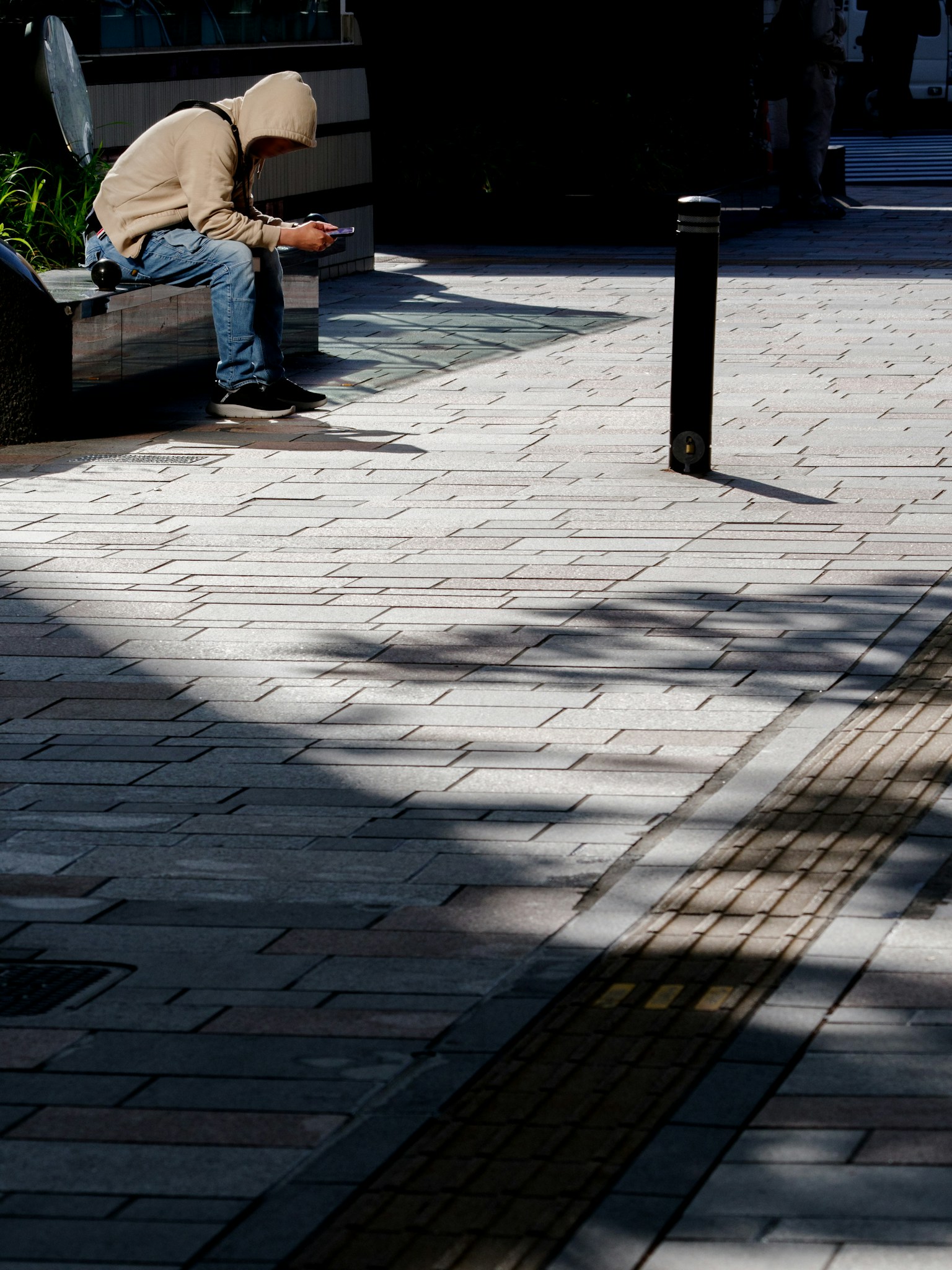 Ein Mann in einer Kapuzenjacke sitzt auf einer Parkbank Schatten auf dem gepflasterten Weg