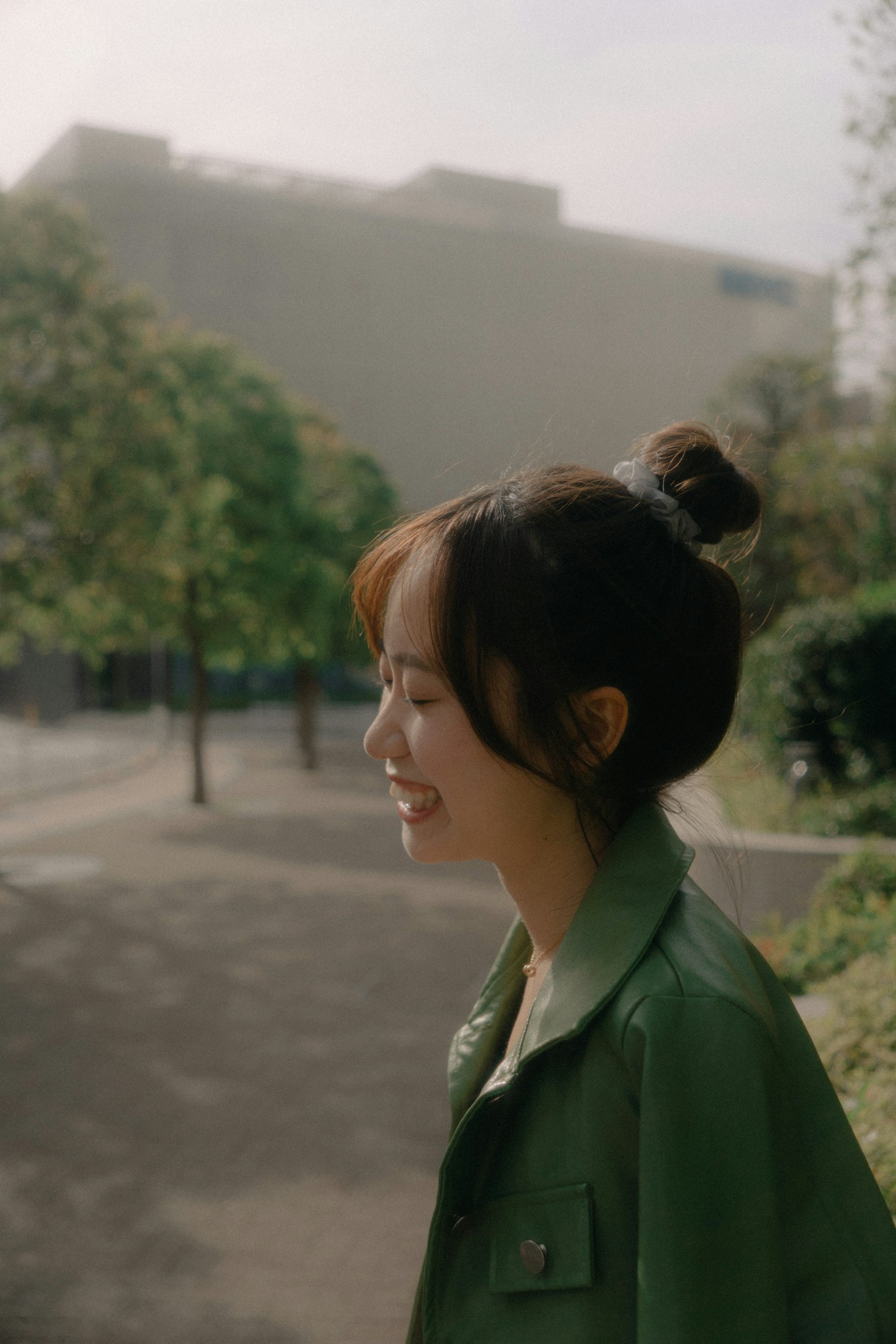 ภาพด้านข้างของผู้หญิงยิ้มในแจ็คเก็ตสีเขียวยืนอยู่ในสวนที่มีต้นไม้และอาคารอยู่เบื้องหลัง