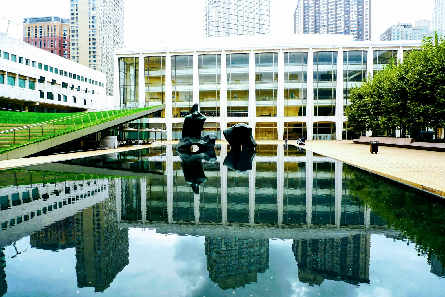 Escena de parque con edificio moderno y esculturas reflejadas en el agua