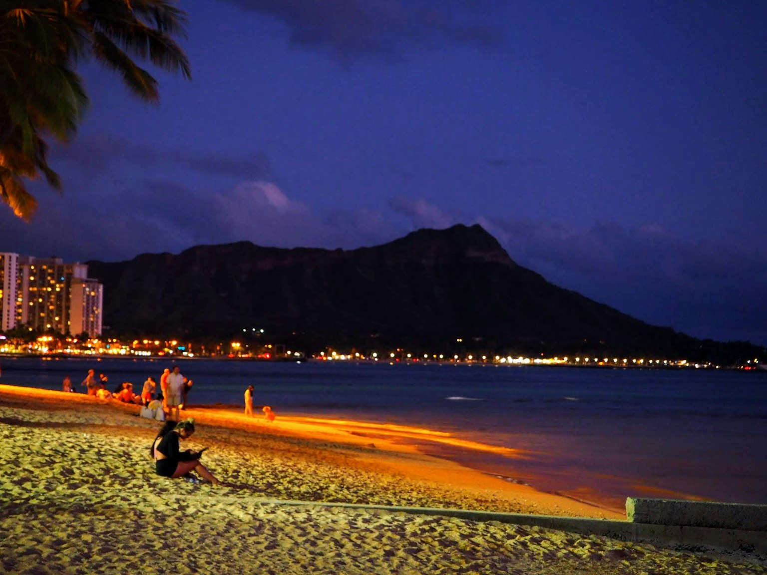 夜晚海滩场景，沙滩上有坐着的人，背景是钻石头山