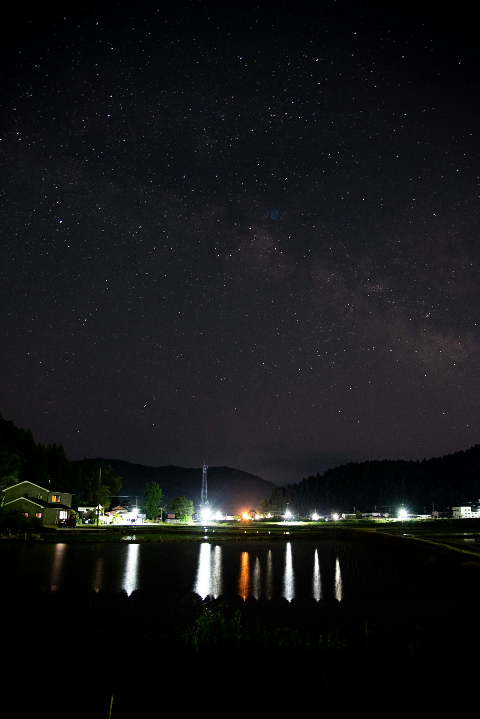 Pemandangan malam dengan langit berbintang yang memantul di danau tenang dikelilingi pegunungan dan cahaya