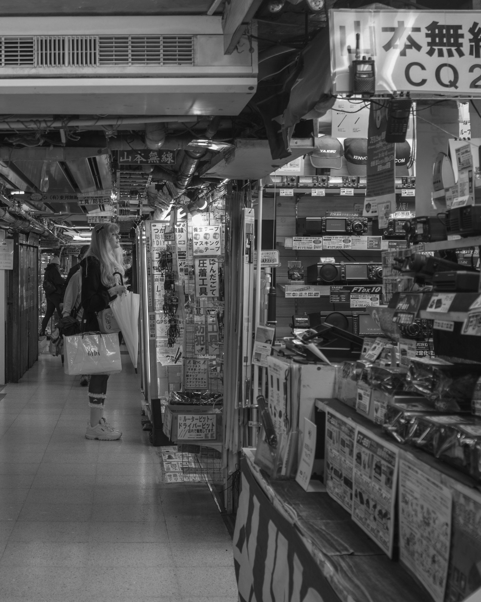 Una mujer de pie en un arcade comercial en blanco y negro con tiendas que exhiben varios productos