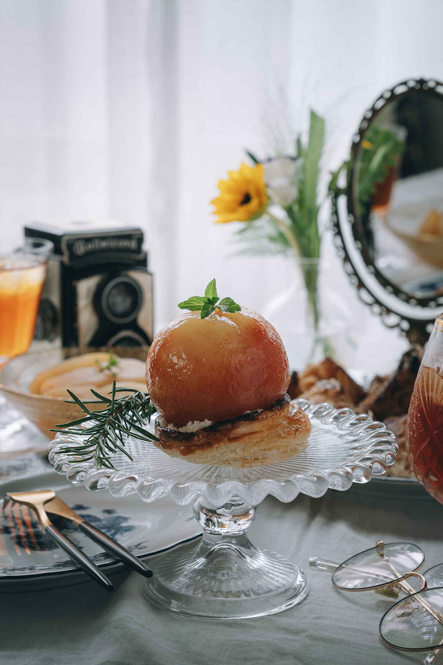 Un dolce di mele splendidamente presentato su un alzata di vetro con fiori e una macchina fotografica vintage sullo sfondo