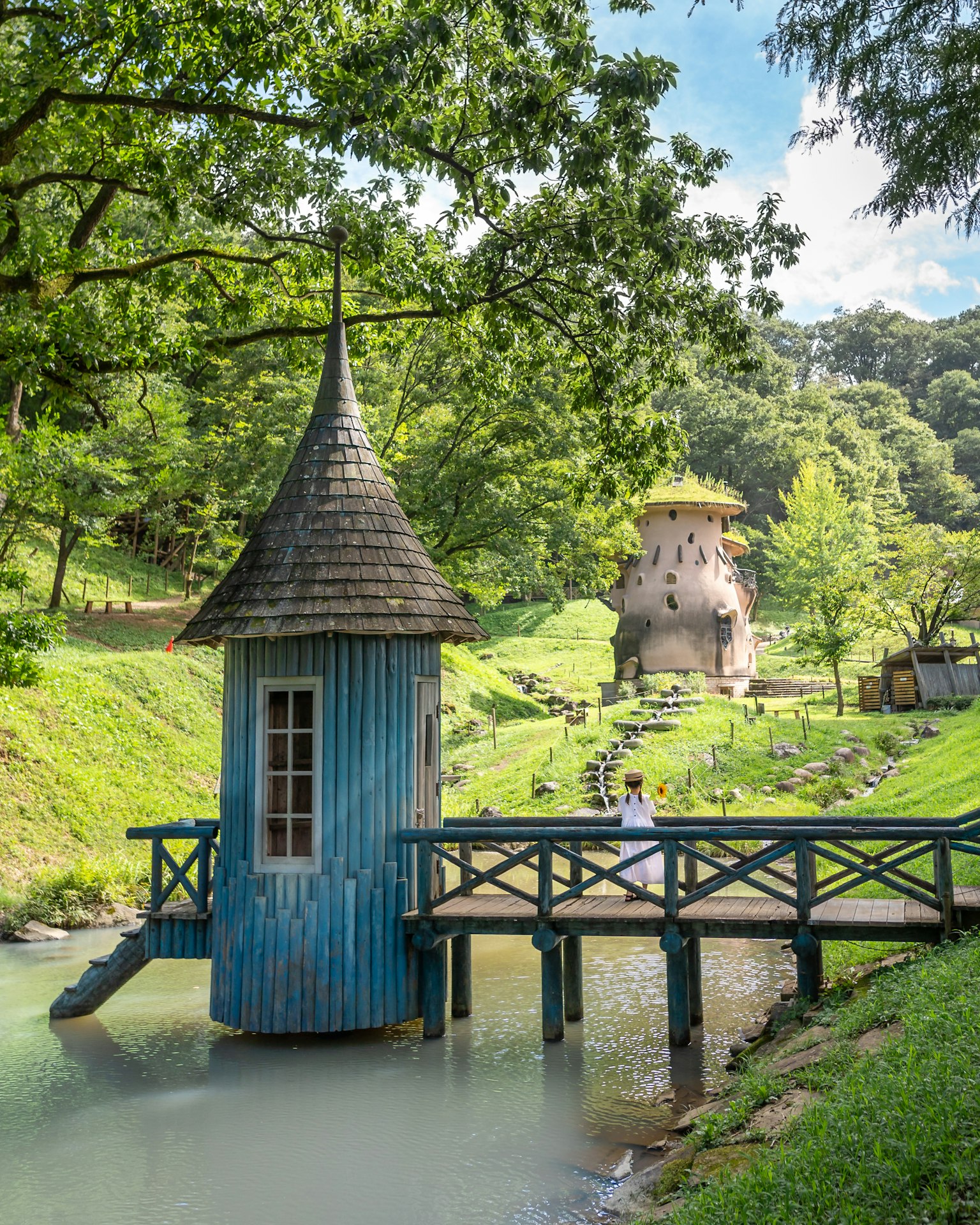 一个风景如画的场景，蓝色的小屋有尖顶，木桥横跨宁静的小溪