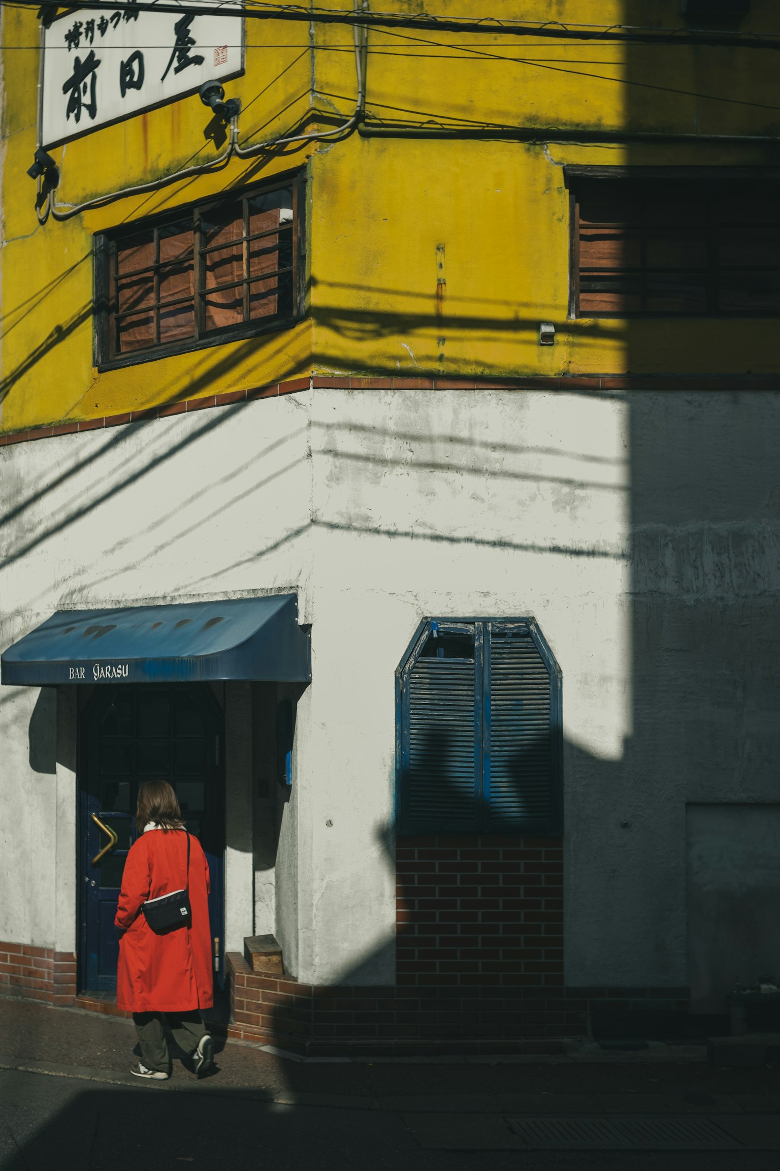 Une femme en manteau rouge se tient au coin d'un bâtiment avec un mur jaune