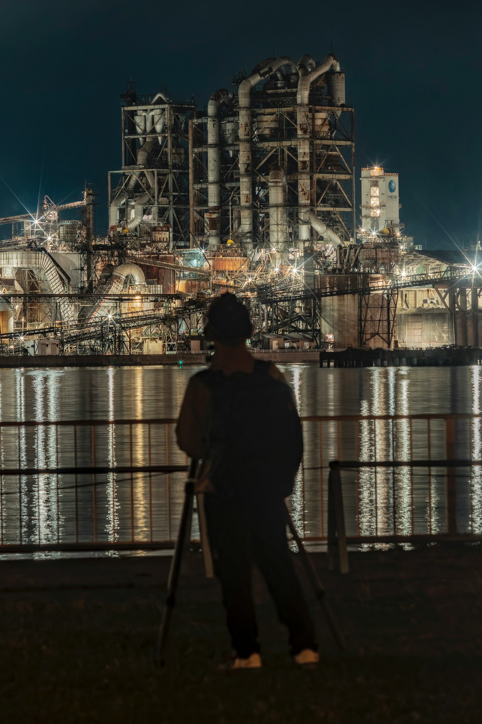 夜の工場の前に立つ人物と水面に映る工場の光