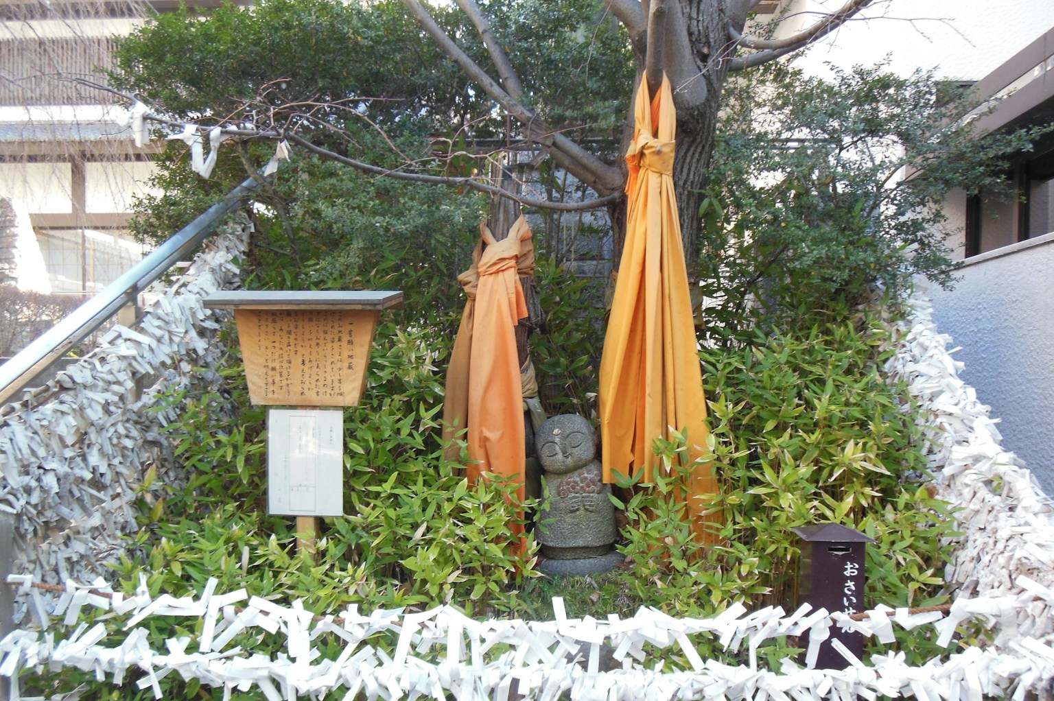 Patung berpakaian oranye dikelilingi tanaman hijau dan kertas omikuji