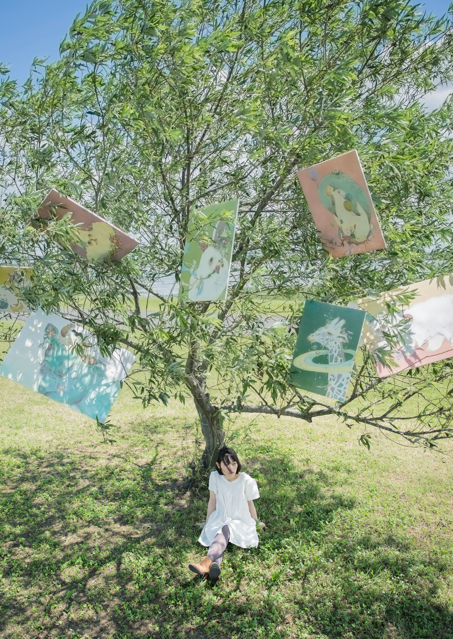 เด็กนั่งอยู่ใต้ต้นไม้สีเขียวที่มีธงภาพสีสันสดใสแขวนอยู่ที่กิ่ง
