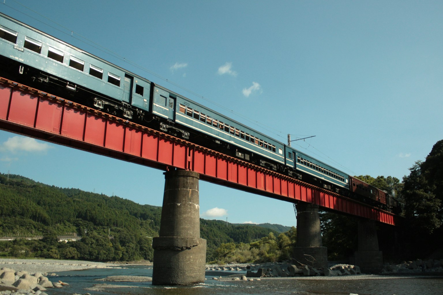 Un tren azul cruzando un puente rojo con un fondo escénico