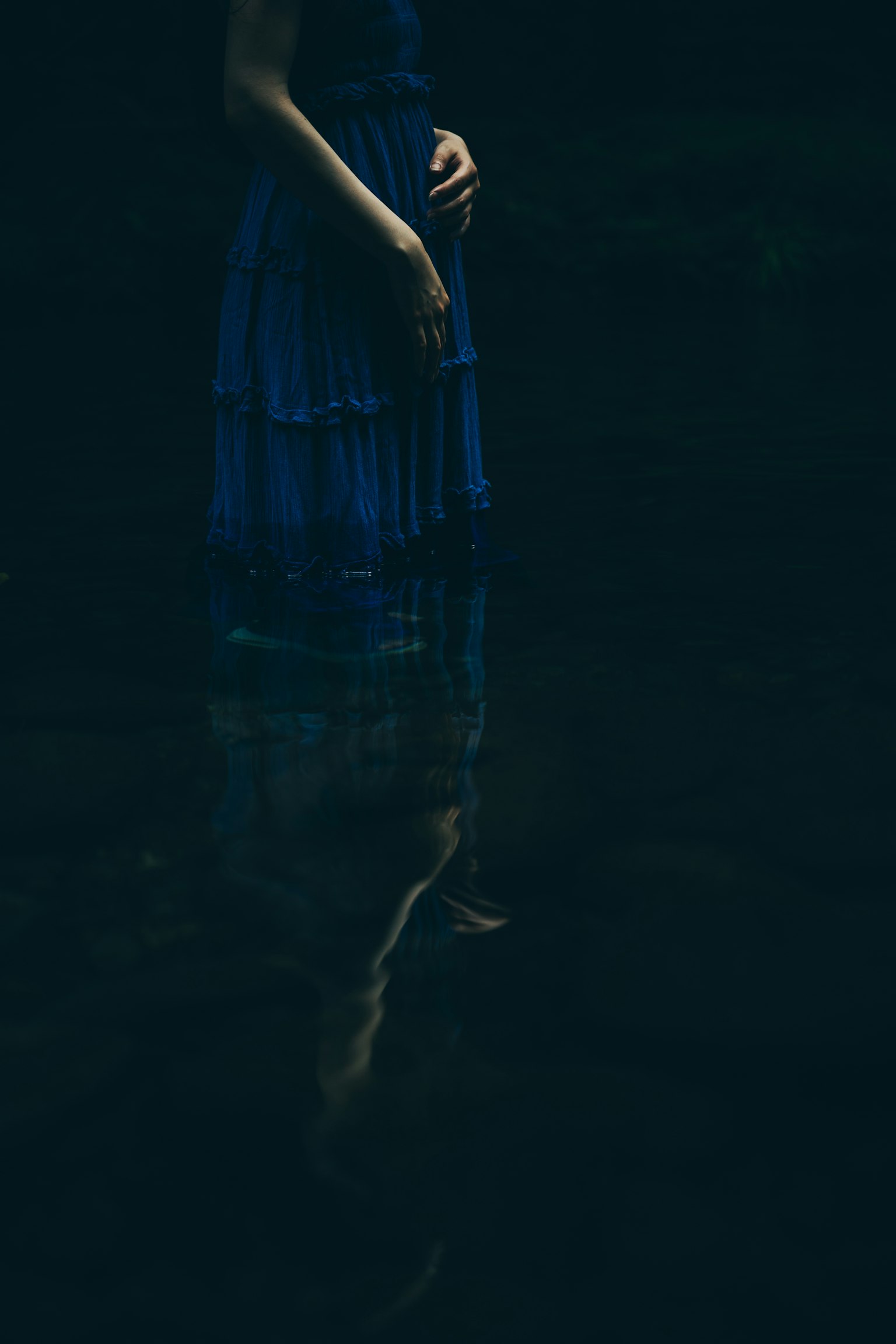 一位穿着蓝色裙子的女性站在水中，背景昏暗，映出倒影