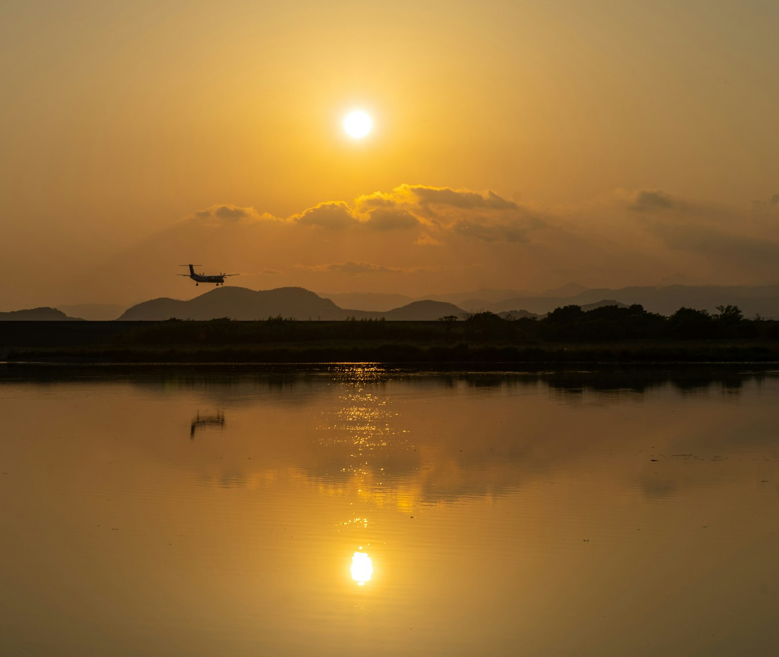 A plane flying over a calm lake reflecting the sunset