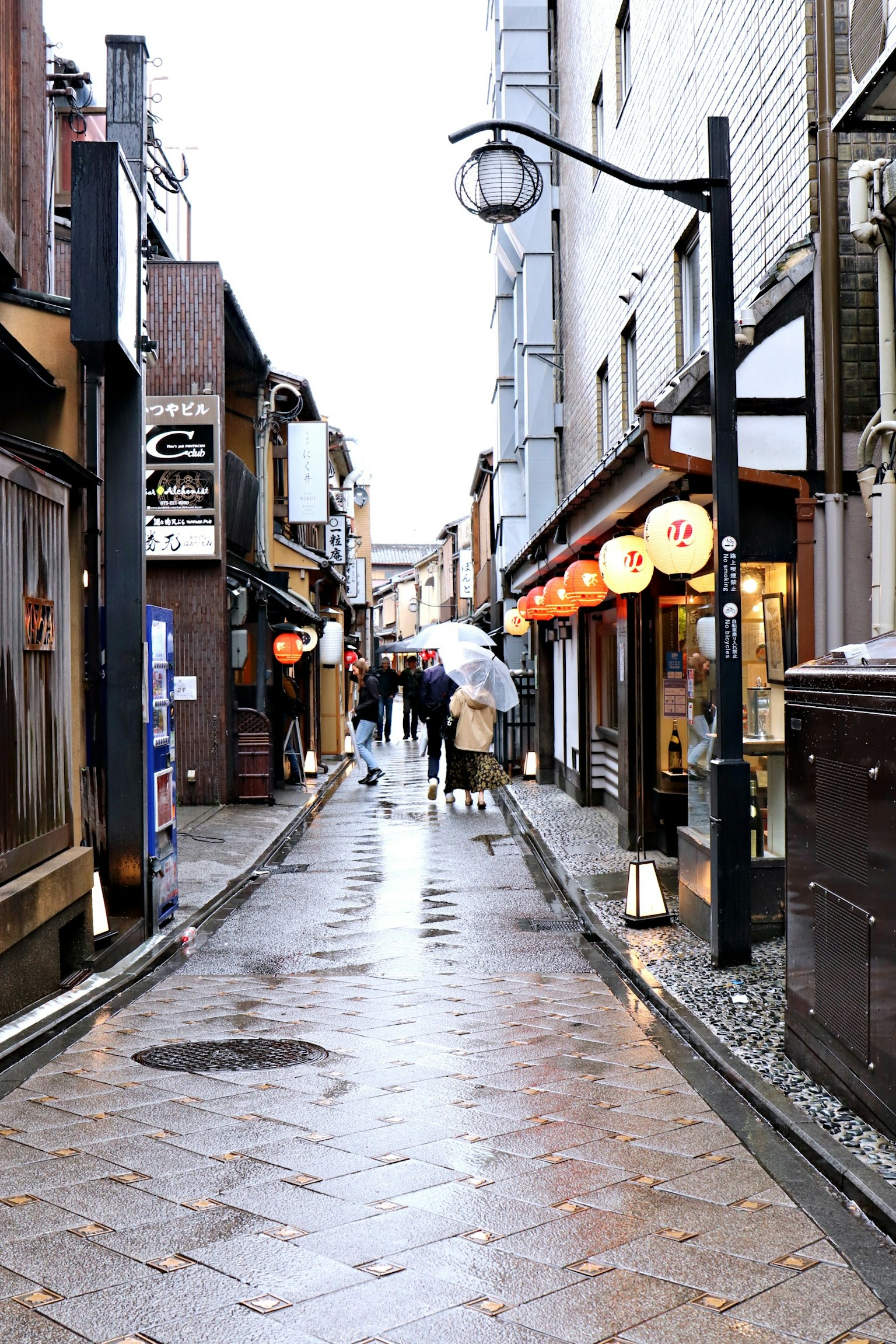 ถนนแคบที่มีร้านค้าและผู้คนถือร่มในฝน