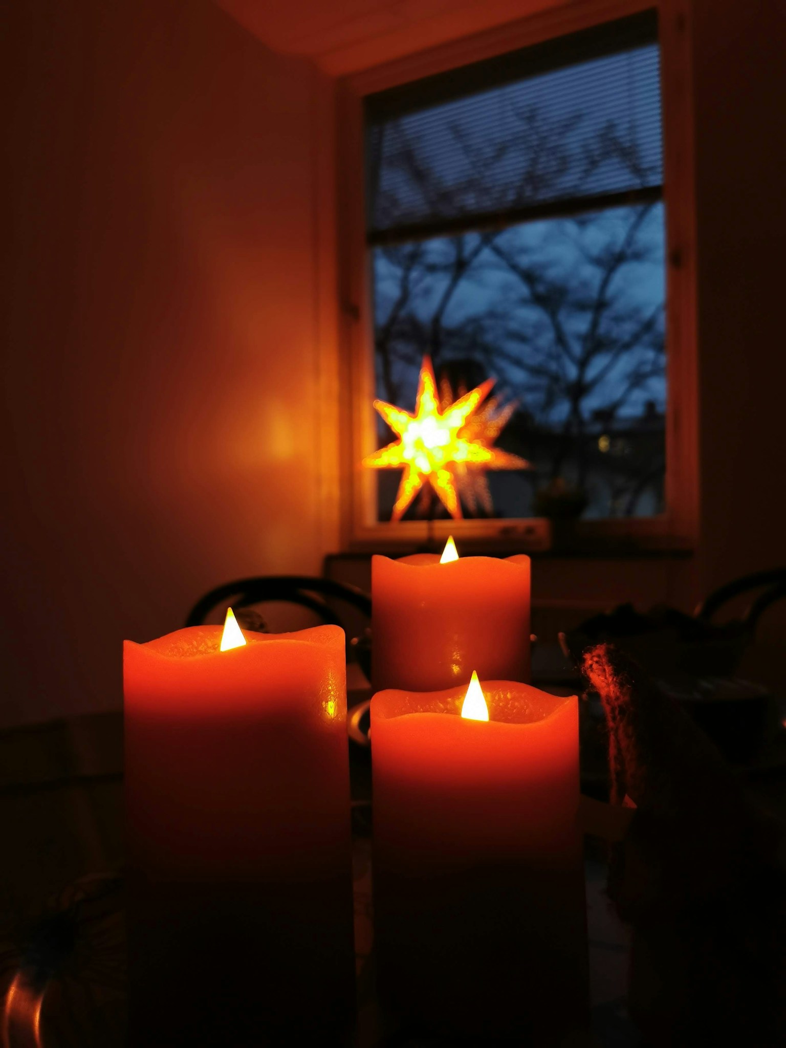 Tre candele arancioni accese in una stanza poco illuminata con una lampada a forma di stella che brilla fuori dalla finestra