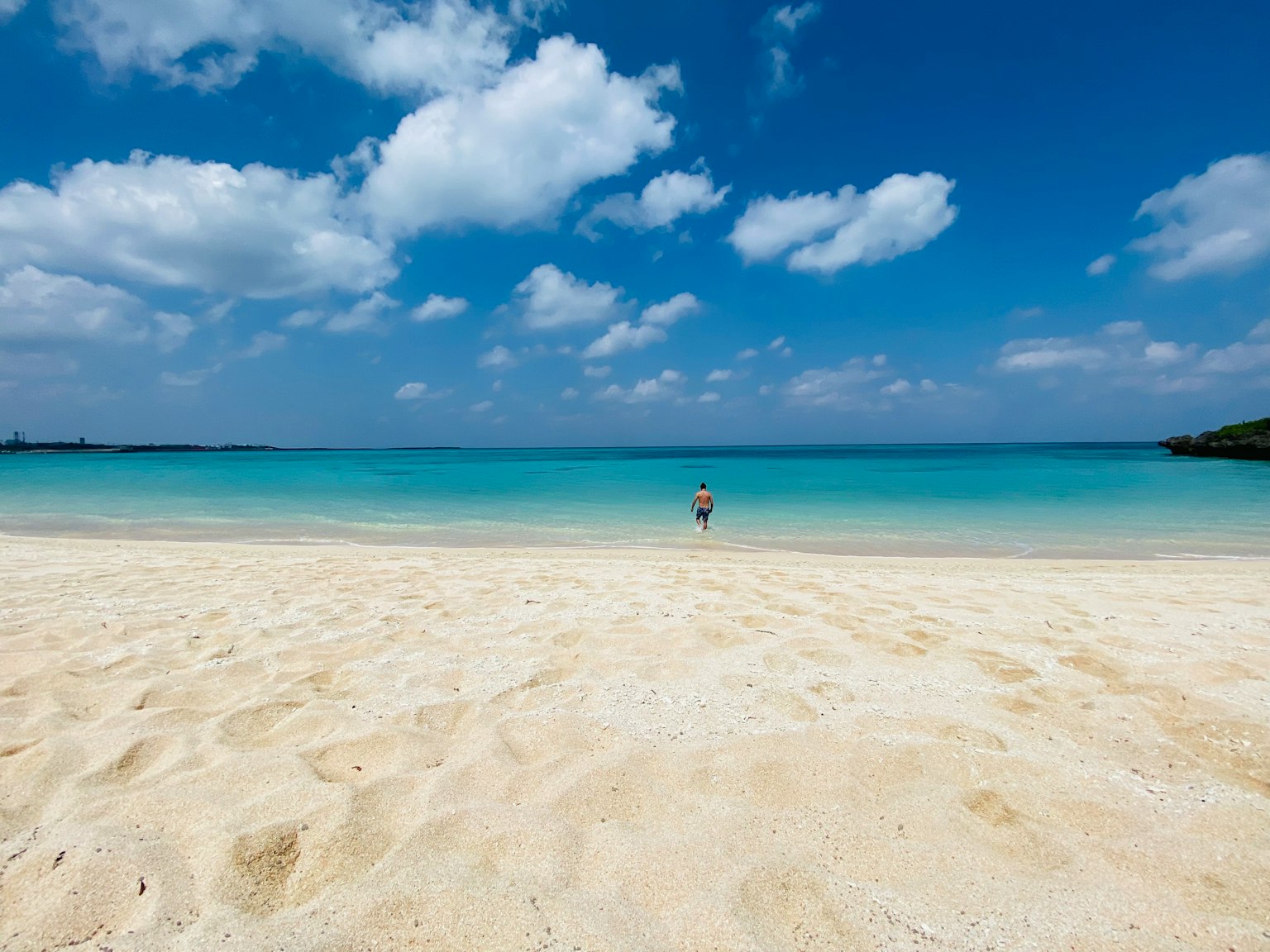Una persona de pie en una playa de arena blanca con océano azul y nubes